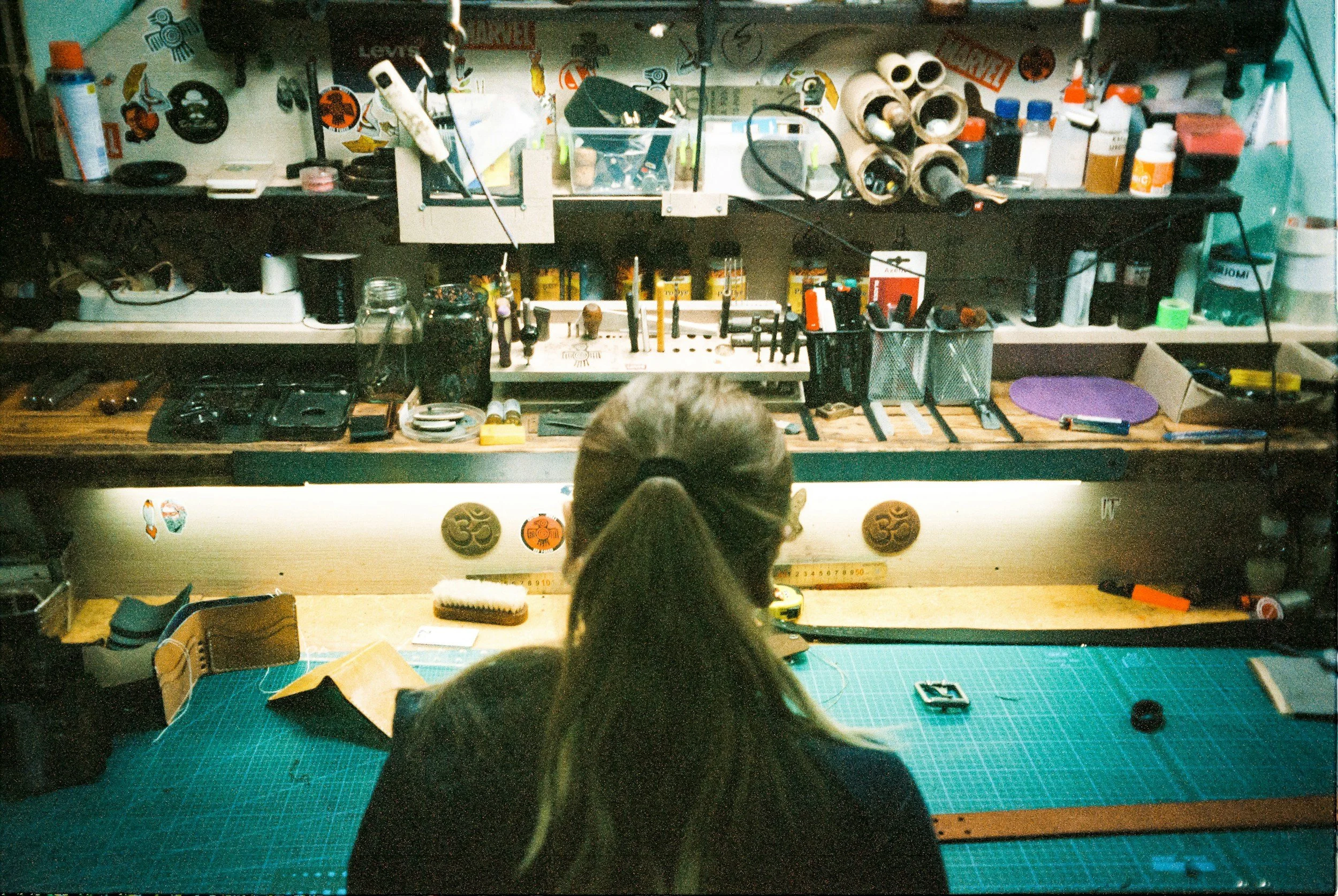 A cluttered workbench with various tools, supplies, and decorations. A person with long hair tied back is working at a green cutting mat, with their back turned, in a workspace filled with small containers, bottles, and stickers on the wall.