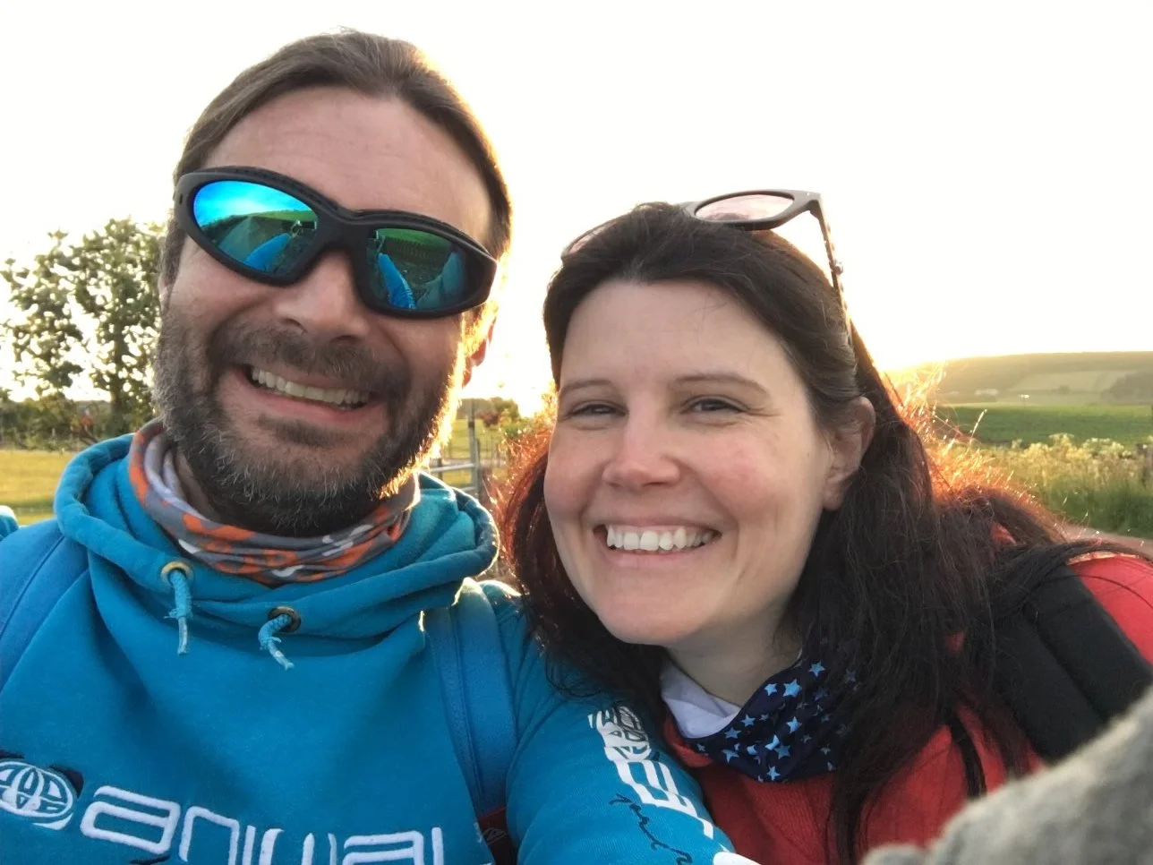A smiling man and woman taking a selfie outdoors during sunset, with green fields and trees in the background.