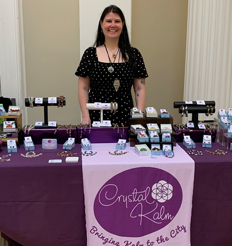 Woman standing behind a jewelry display table with various necklaces, bracelets, and jewelry boxes, at a craft fair or market.