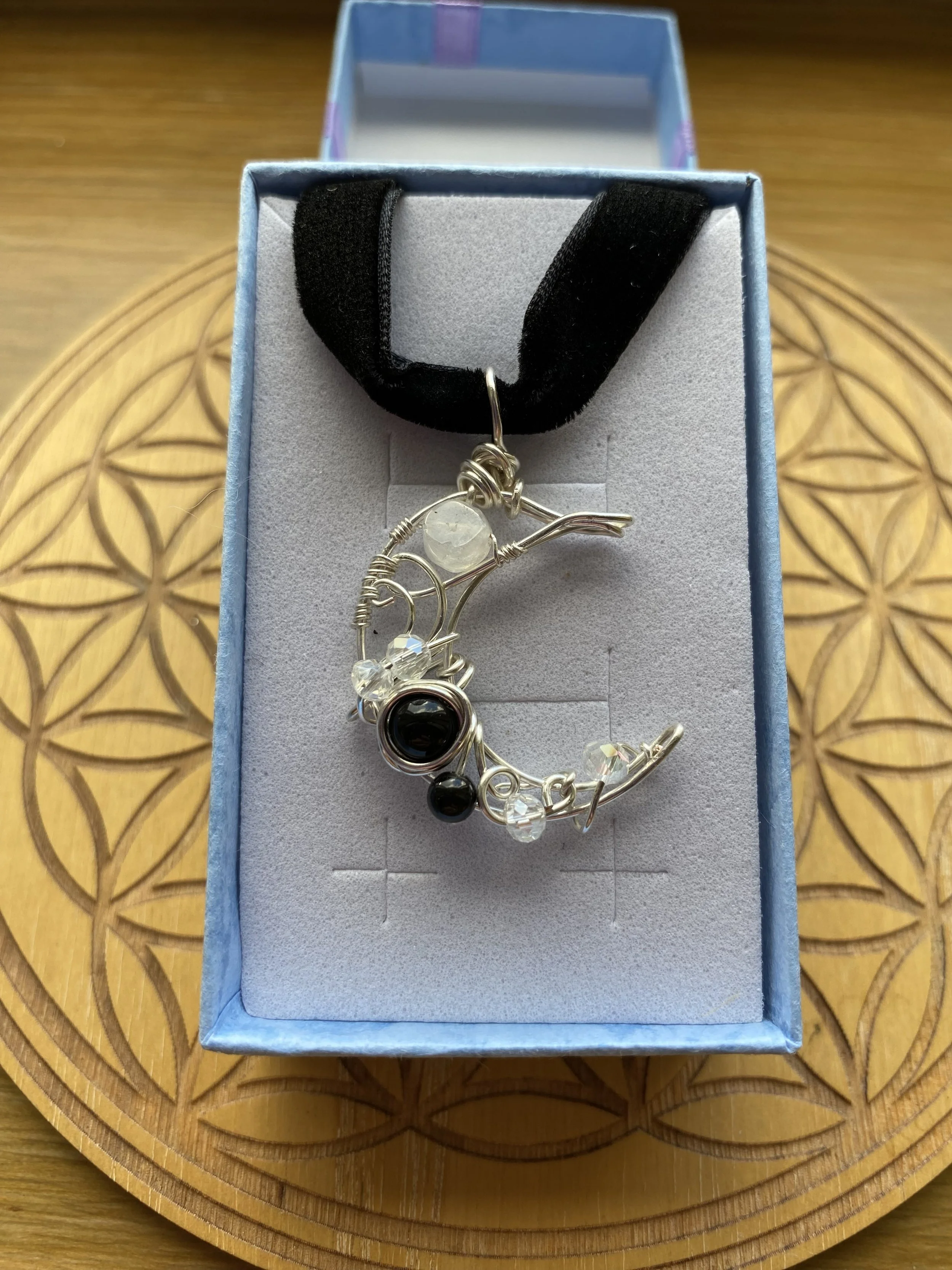 A moon necklace with Black Obsidian and clear beads, featuring a silver wire design, placed in a light blue jewellery box on a wooden table with a circular pattern.