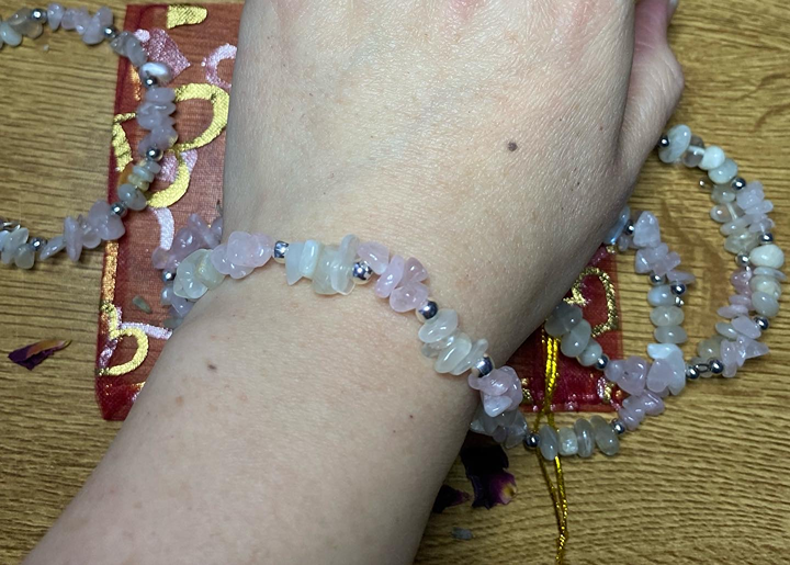 A person's wrist wearing a beaded bracelet with Rose Quartz and Moonstone crystal chips, resting on a wooden surface with a decorative red bag and a beaded necklace nearby.