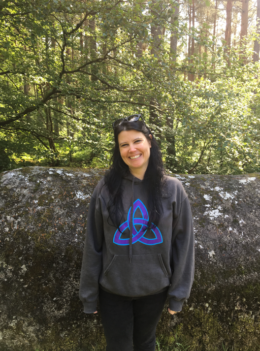 A woman with long dark hair, smiling and wearing sunglasses on her head, stands in front of a large mossy rock in a lush forest with sunlight filtering through the trees. She is wearing a dark gray hoodie with a blue geometric design on the front.