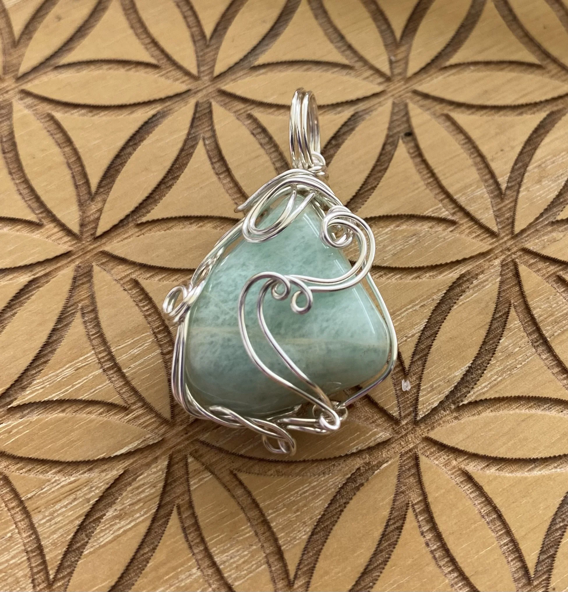 An Amazonite crystal wrapped in silver wire with intricate swirls, resting on a wooden surface with a carved floral pattern.