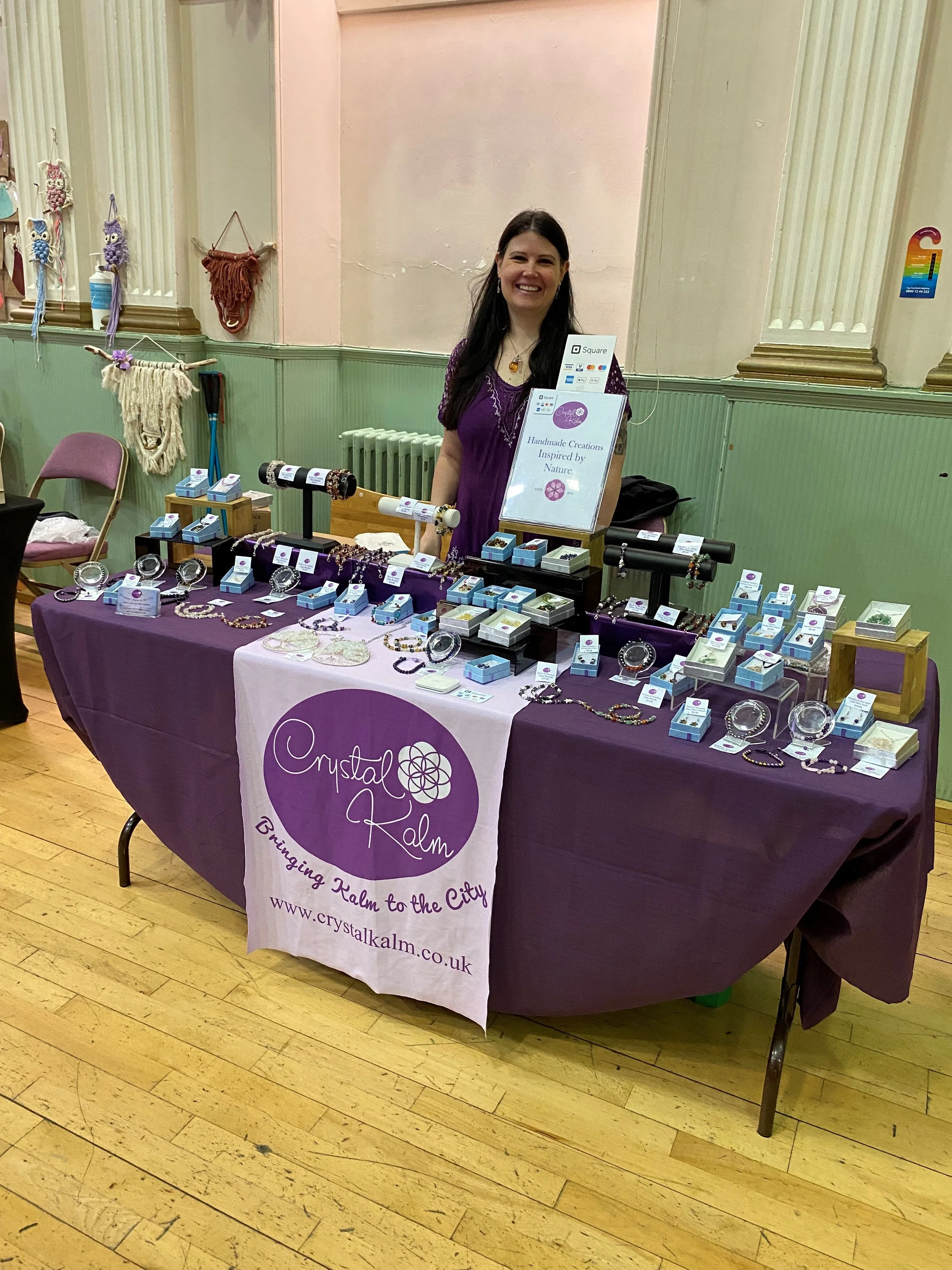 A smiling woman standing behind a table with handmade jewelry at a craft fair. The table is covered with a purple tablecloth and has various jewelry displayed in boxes and on stands. The booth has a sign that reads 'Crystal Kalm' and a banner with the same name and website URL.