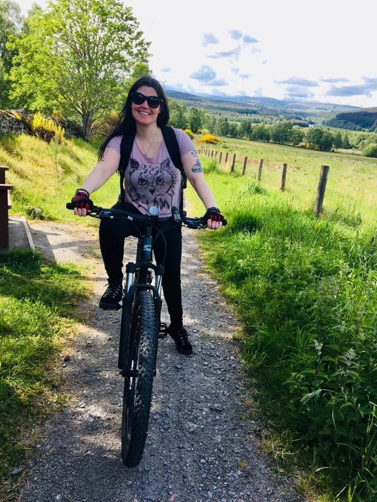 A woman with long dark hair, wearing sunglasses, a pink owl t-shirt, black pants, and gloves, posing with a mountain bike on a dirt trail in a green rural landscape with trees, grass, fences, and cloudy sky.