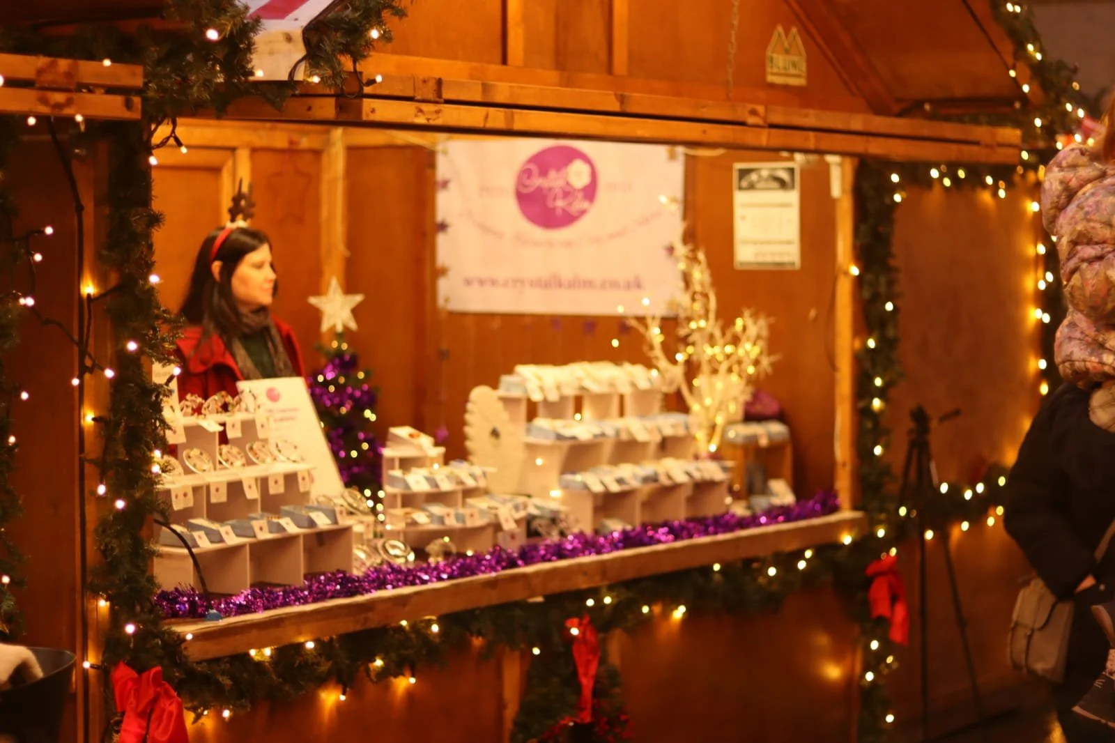 A woman standing behind a Christmas market stall decorated with purple tinsel and string lights. The stall displays various jewelry items, with a decorated small Christmas tree and a sign in the background. The stall is made of wood and has a festive, holiday-themed setting.