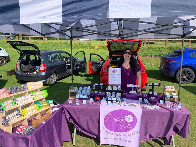 Woman standing behind her jewelry stall at an outdoor market under a blue and white canopy, with cars parked on a grassy field in the background.