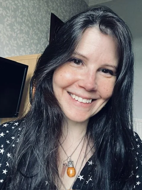 A woman with long black hair smiling in a room with patterned wallpaper, a black TV, and a wooden piece of furniture.
