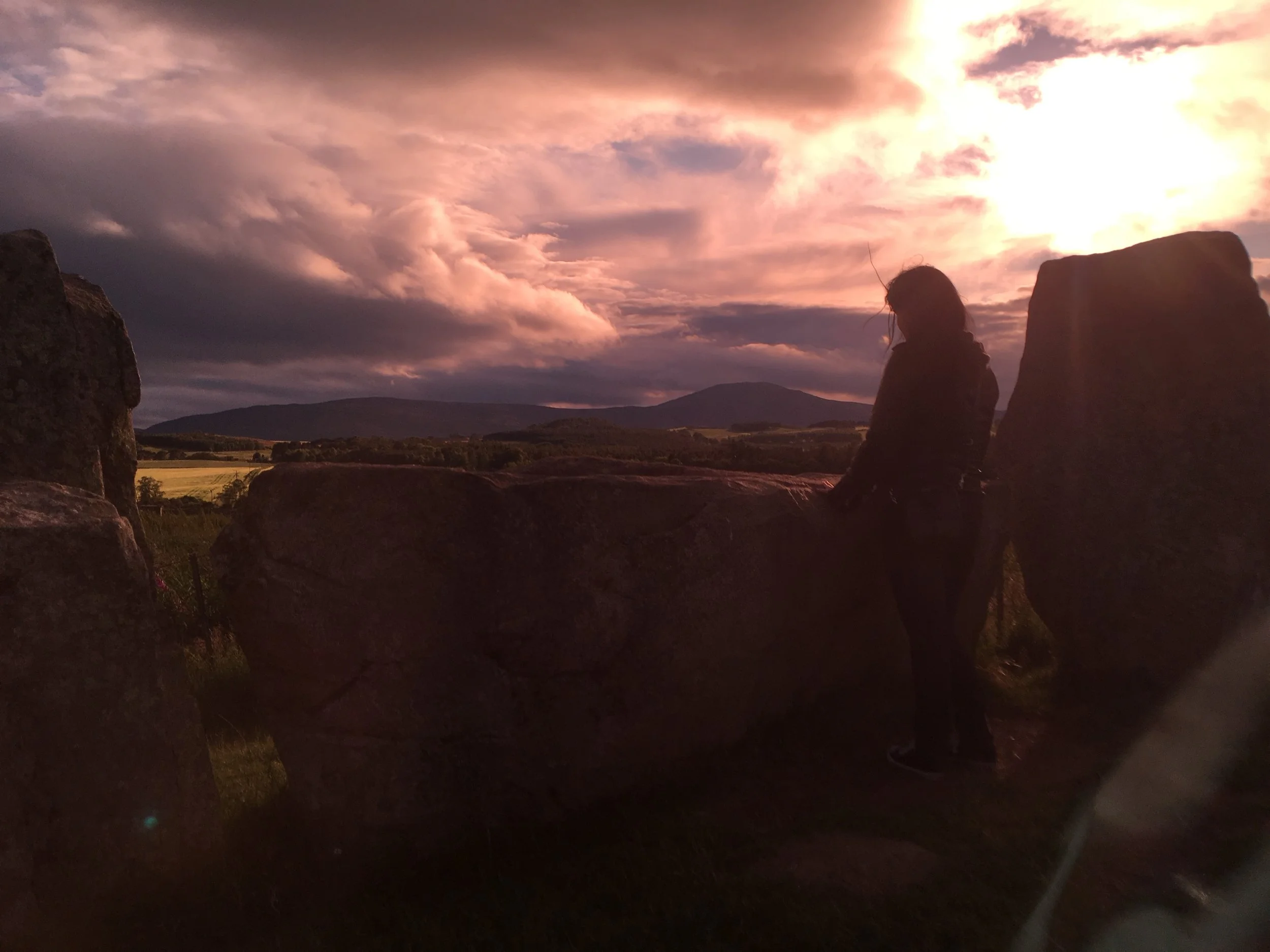 A person sitting on a rock formation at sunset with cloudy sky and rolling hills in the background.