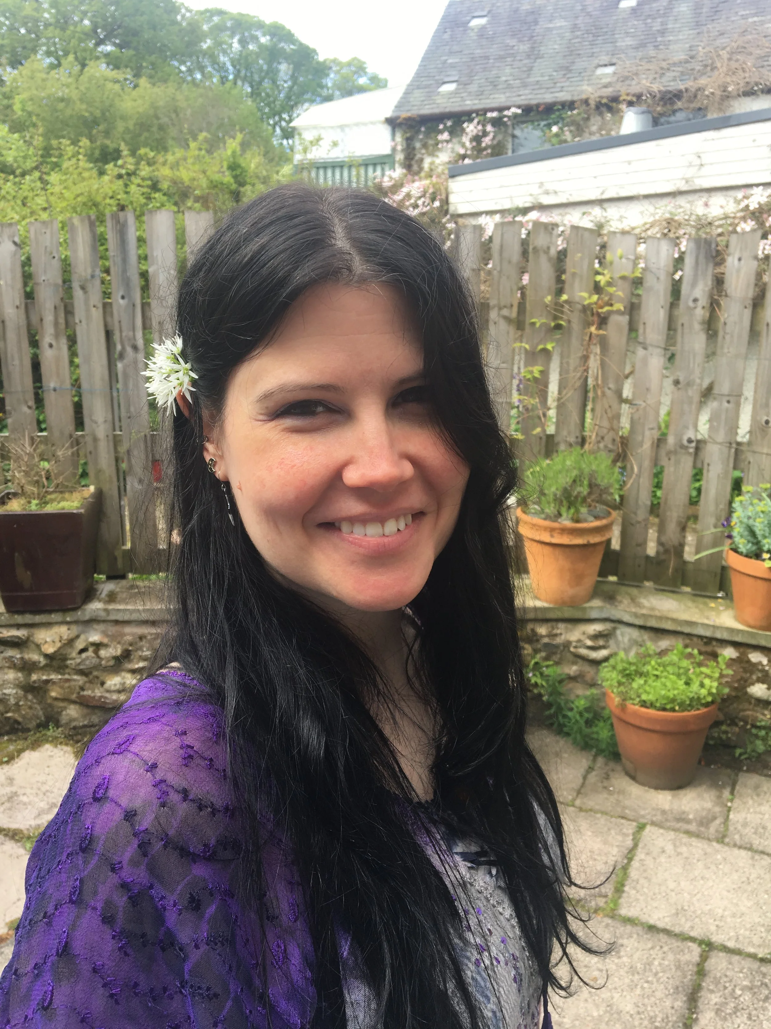 A smiling woman with dark hair and a white flower in her hair, standing outdoors in a garden with potted plants and a wooden fence in the background.