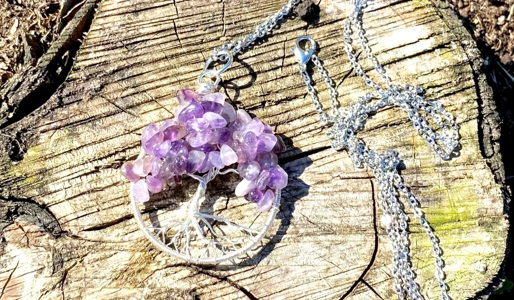 A silver wire tree of life jewellery pendant with Amethyst gemstone chips on a rustic wooden surface, with a flowing silver chain beside it.