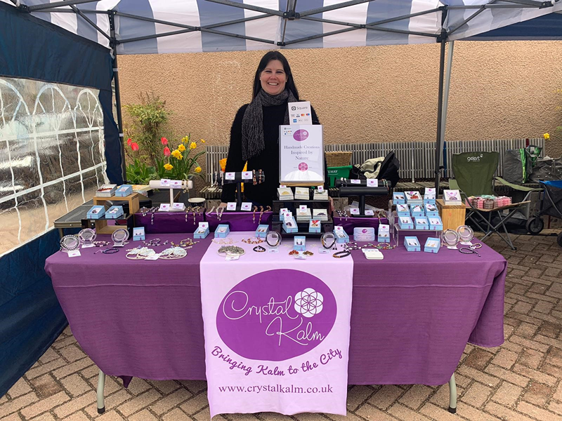 Woman smiling at a jewelry booth with a purple tablecloth displaying jewelry items, under a canopy at an outdoor market