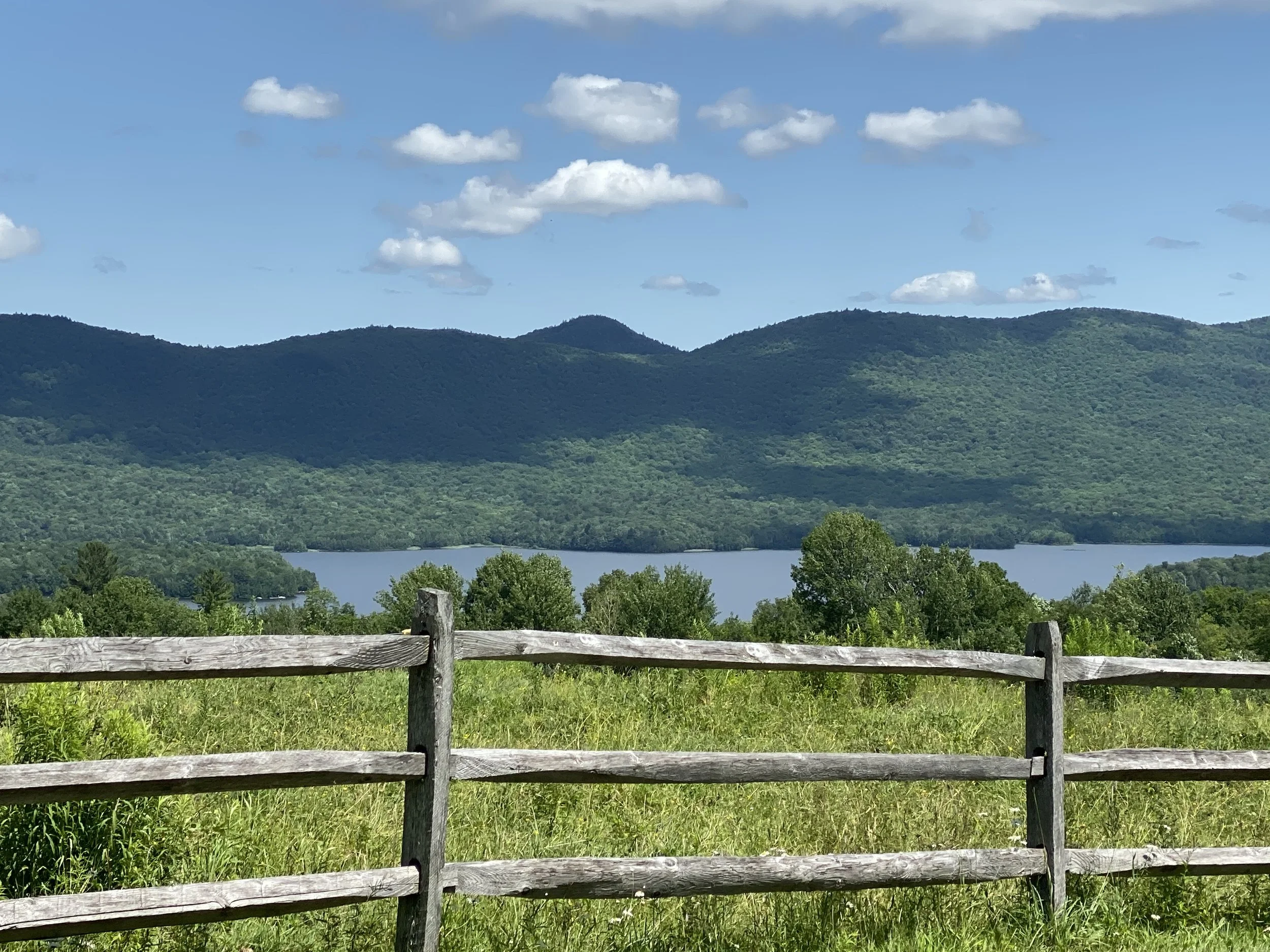 Scenic view of a lake surrounded by green hills and mountains under a blue sky with scattered clouds, seen from behind a wooden fence.