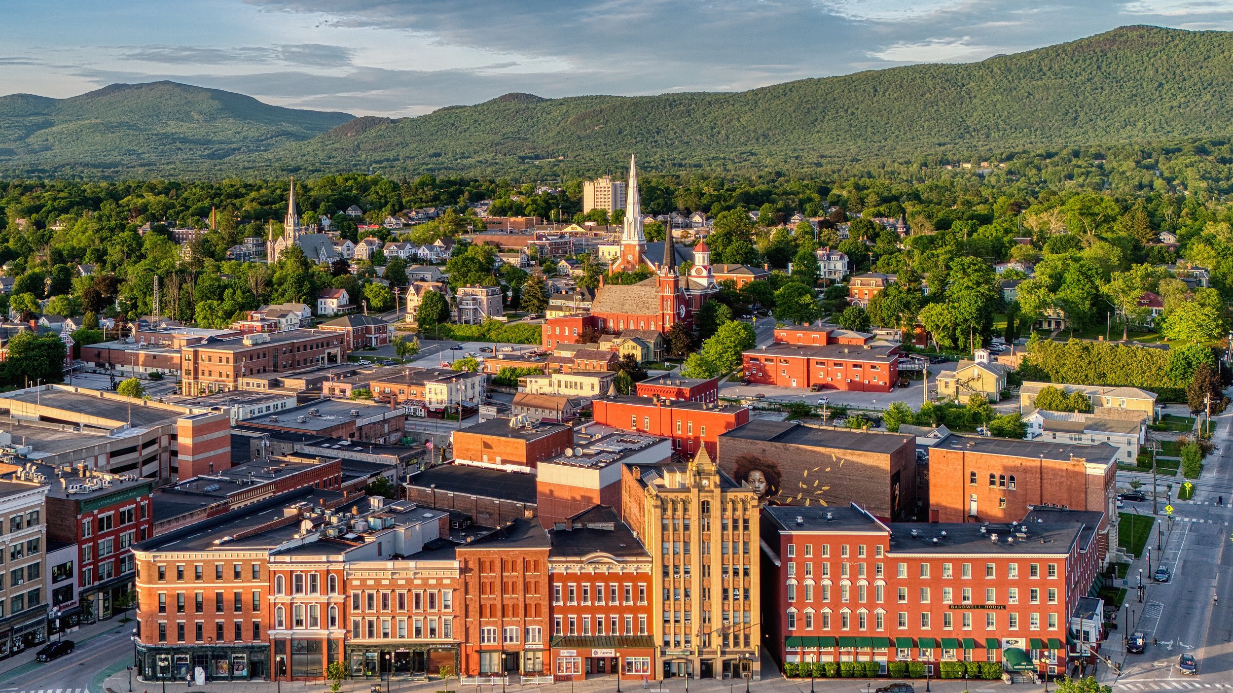 Aerial view of a small downtown with red brick buildings, church steeples, and green trees with a mountain range in the background.