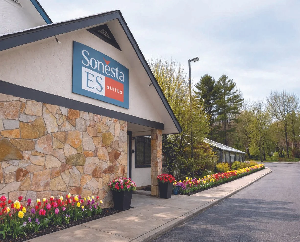 Exterior view of Sonesta ES Suites hotel with colorful flower beds, stone facade, and a parking lot under an overcast sky.