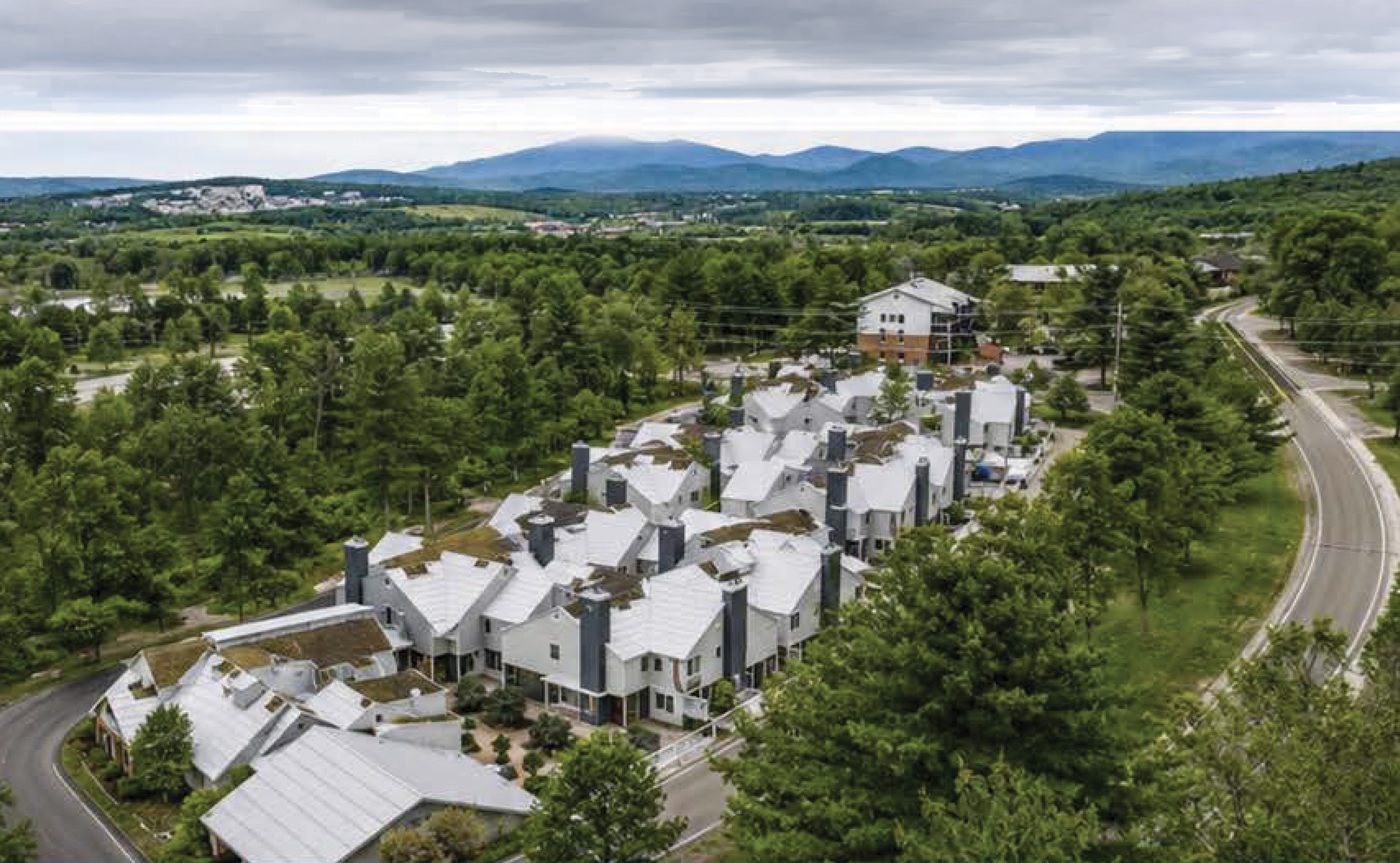 Aerial view of a residential area with white buildings and gray rooftops surrounded by lush green trees, with mountains in the distance under a cloudy sky.