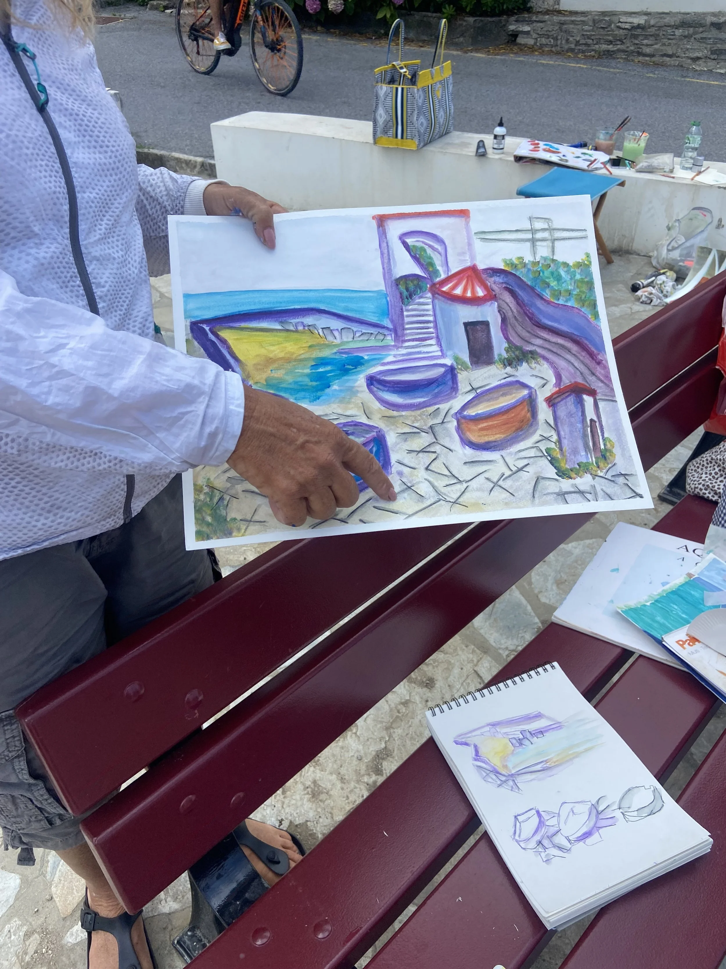 Un homme tient une tableau de peinture représentant une scène colorée avec une vue sur la mer, un escalier, et des boats, lors d'une session artistique en plein air.