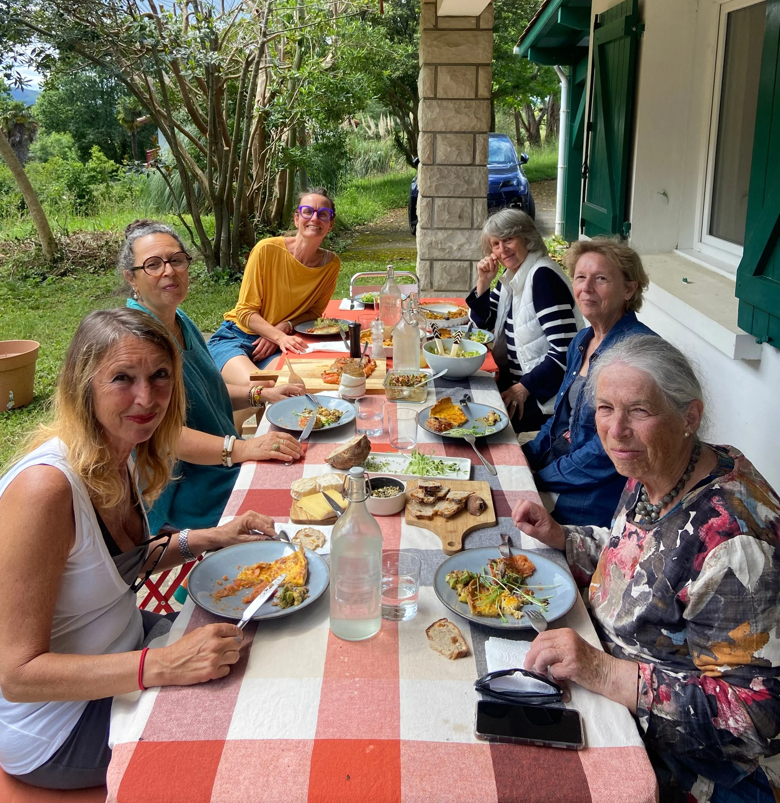 Groupe de personnes assises autour d'une table en plein air, partageant un repas convivial sous un abri, avec de la verdure en arrière-plan.