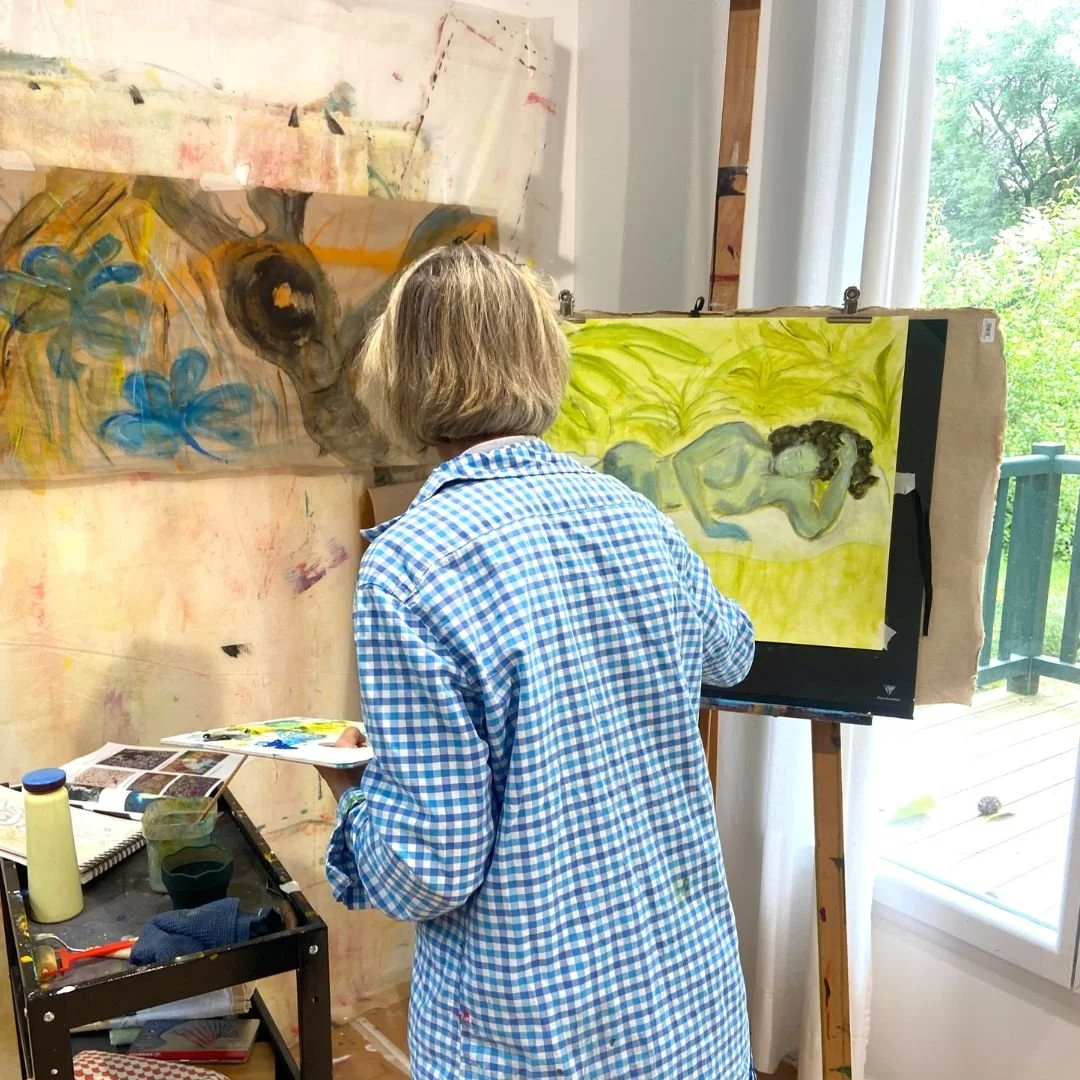 Une artiste peint un tableau dans un atelier, vue de dos, autour de toiles, peintures et outils, avec une grande fenêtre laissant entrer la lumière
