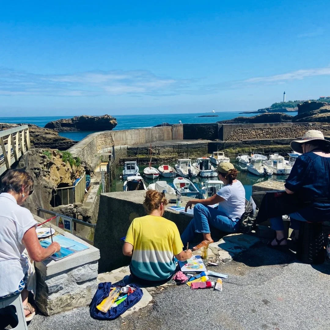 Groupe de personnes dessinant et peignant au bord d'un port de pêche avec des bateaux, la mer et un phare en arrière-plan.