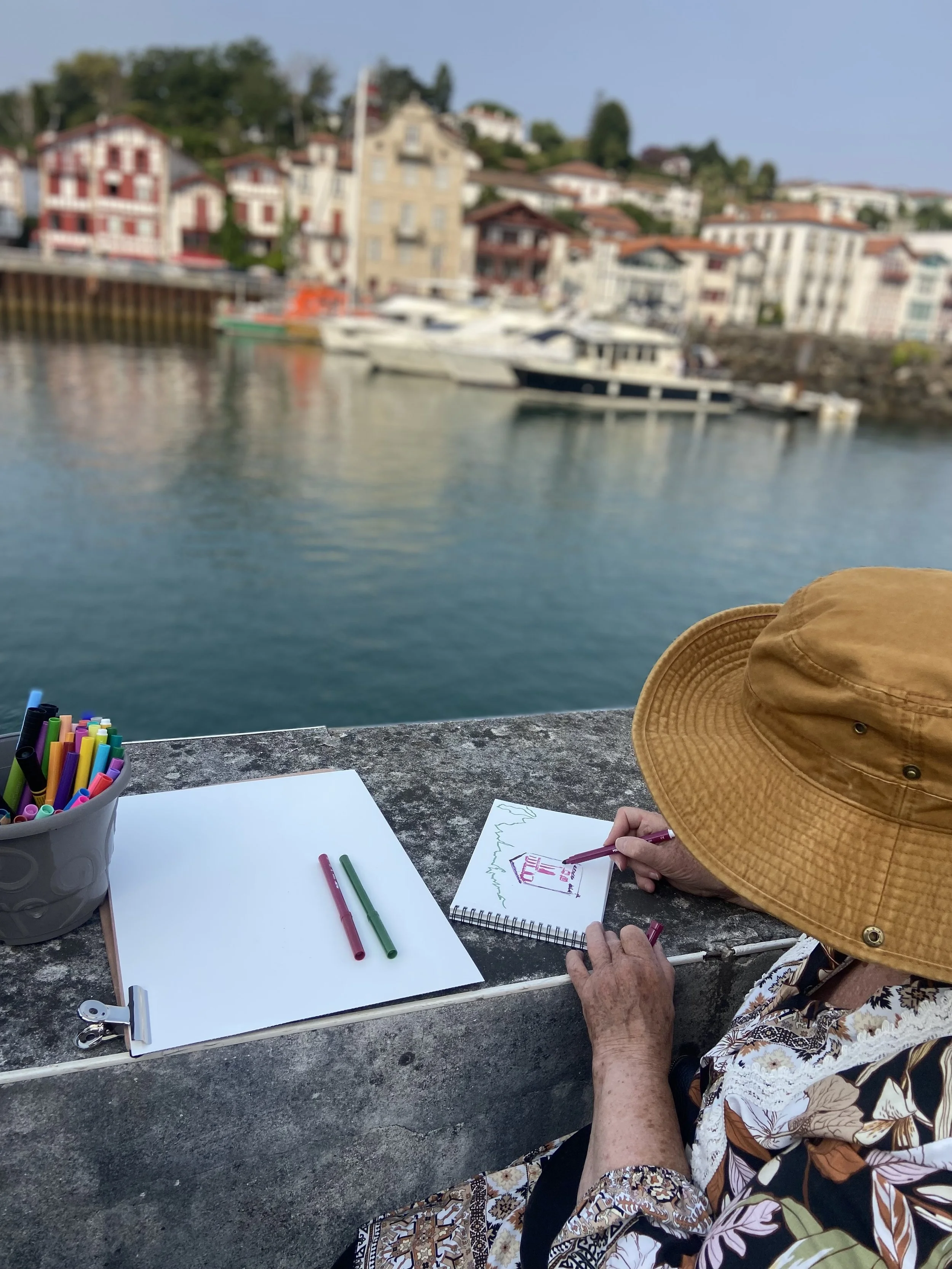 Une personne âgée portant un chapeau beige et vêtue d'une chemise à motifs floraux, assise à une table en faisant du dessin ou de l'écriture, avec un paysage urbain en arrière-plan incluant des maisons colorées et des bateaux dans l'eau.