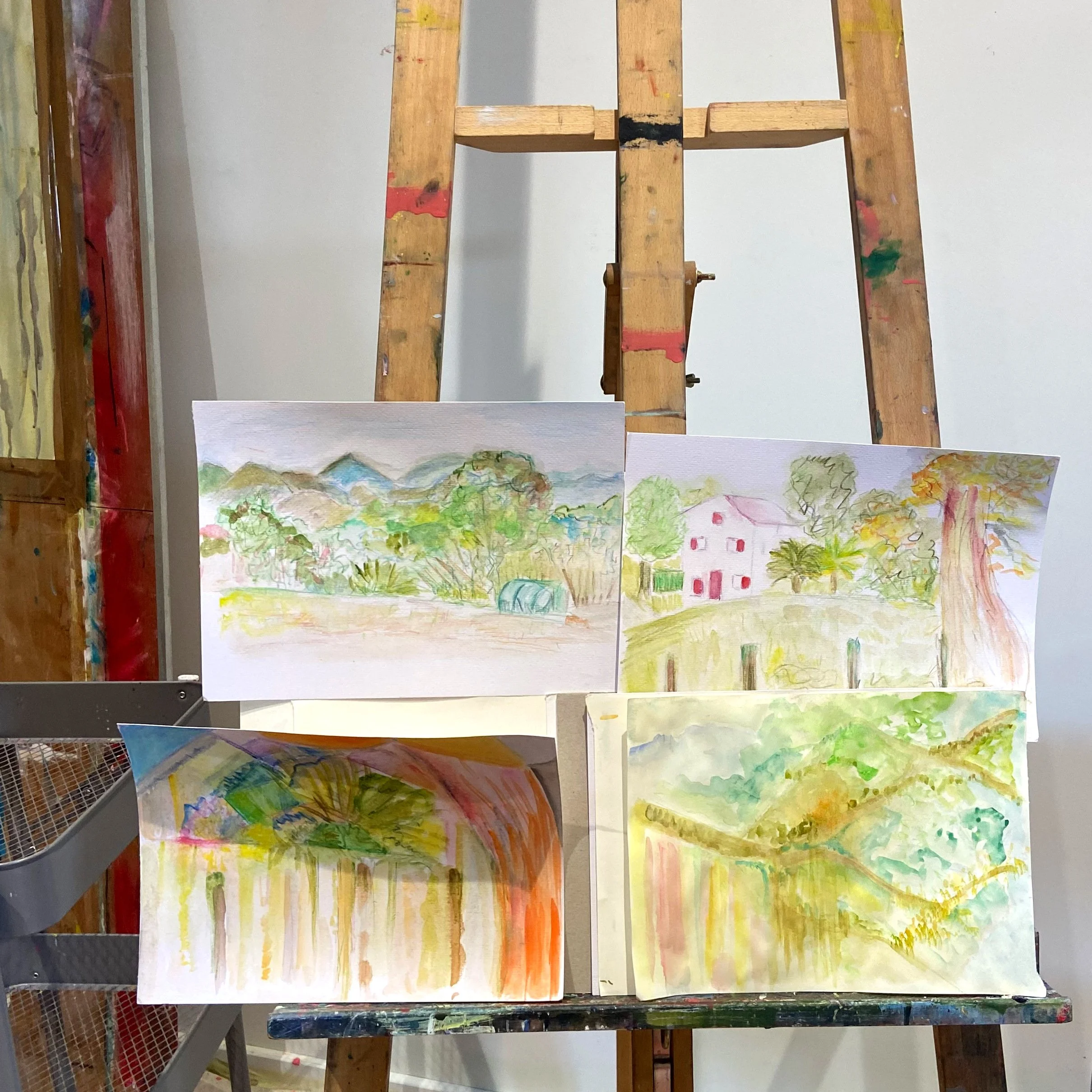 Tableau de plusieurs peintures à l'aquarelle représentant des paysages ruraux avec des maisons, arbres et montagnes, posées sur une table avec un chevalet de peinture en bois.