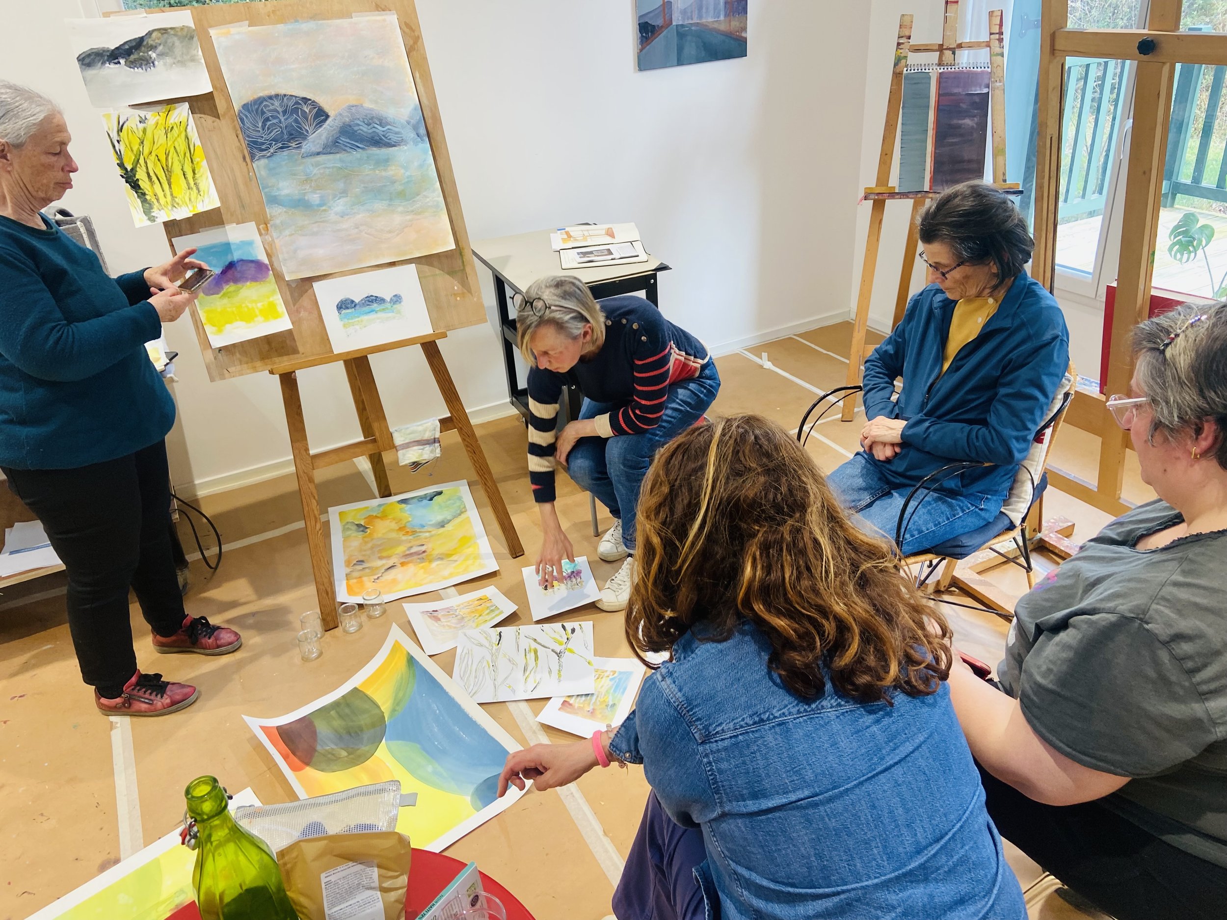 Groupe de personnes dans un atelier de peinture, entouré de tableaux colorés et d'échantillons de peinture, certains regardant et d'autres manipulant des œuvres d'art ou des matériaux.