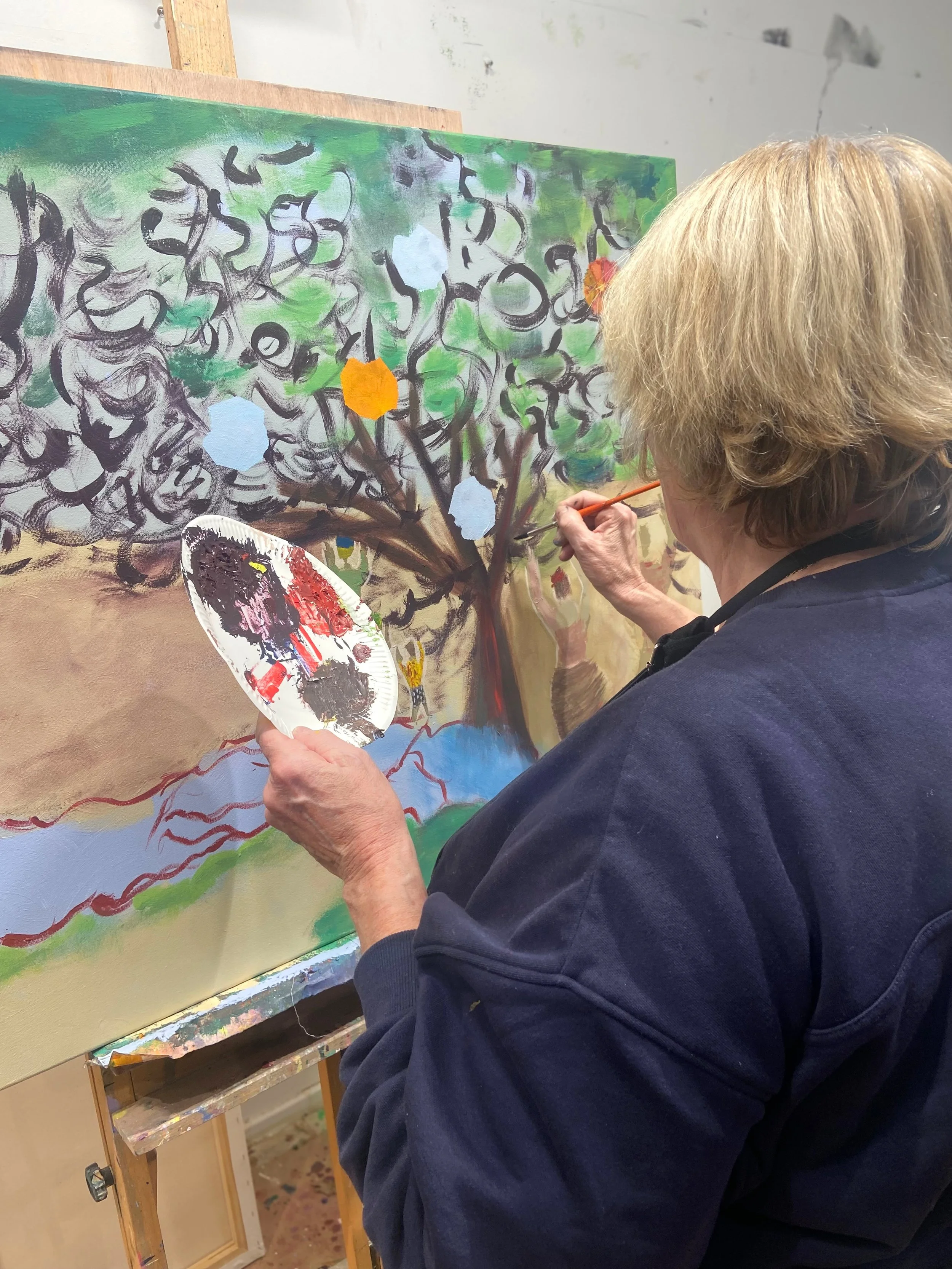 Une femme âgée peint un tableau représentant un arbre avec des branches noires, un fond vert et quelques formes colorées comme des feuilles ou des fleurs.