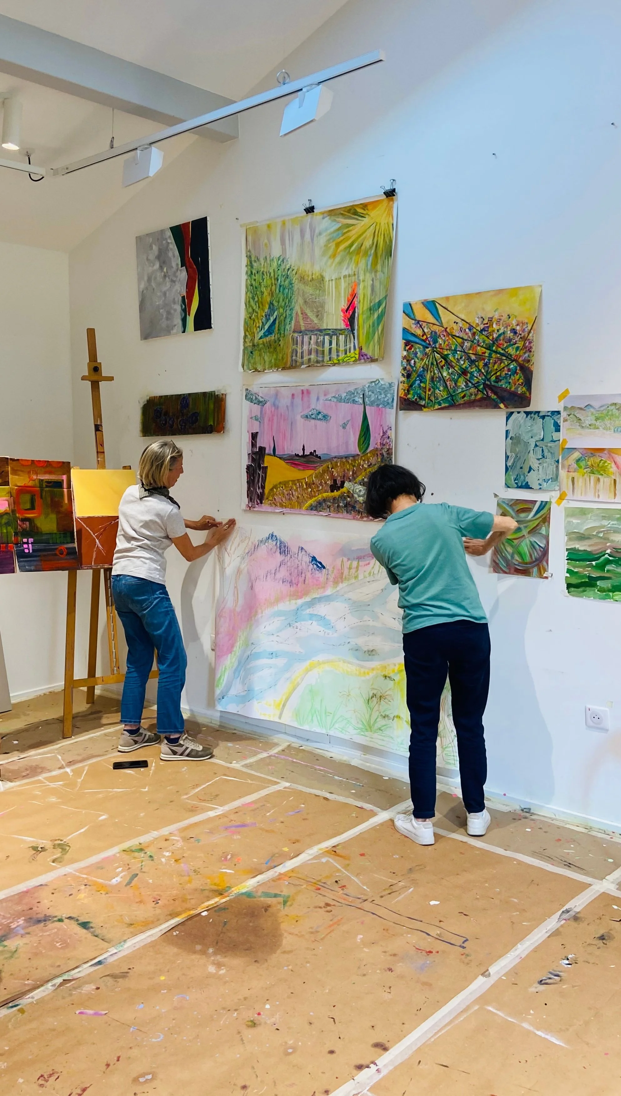 Deux femmes travaillent sur des œuvres d'art dans un atelier de peinture ; les tableaux sont accrochés au mur et la surface de travail est couverte de peinture