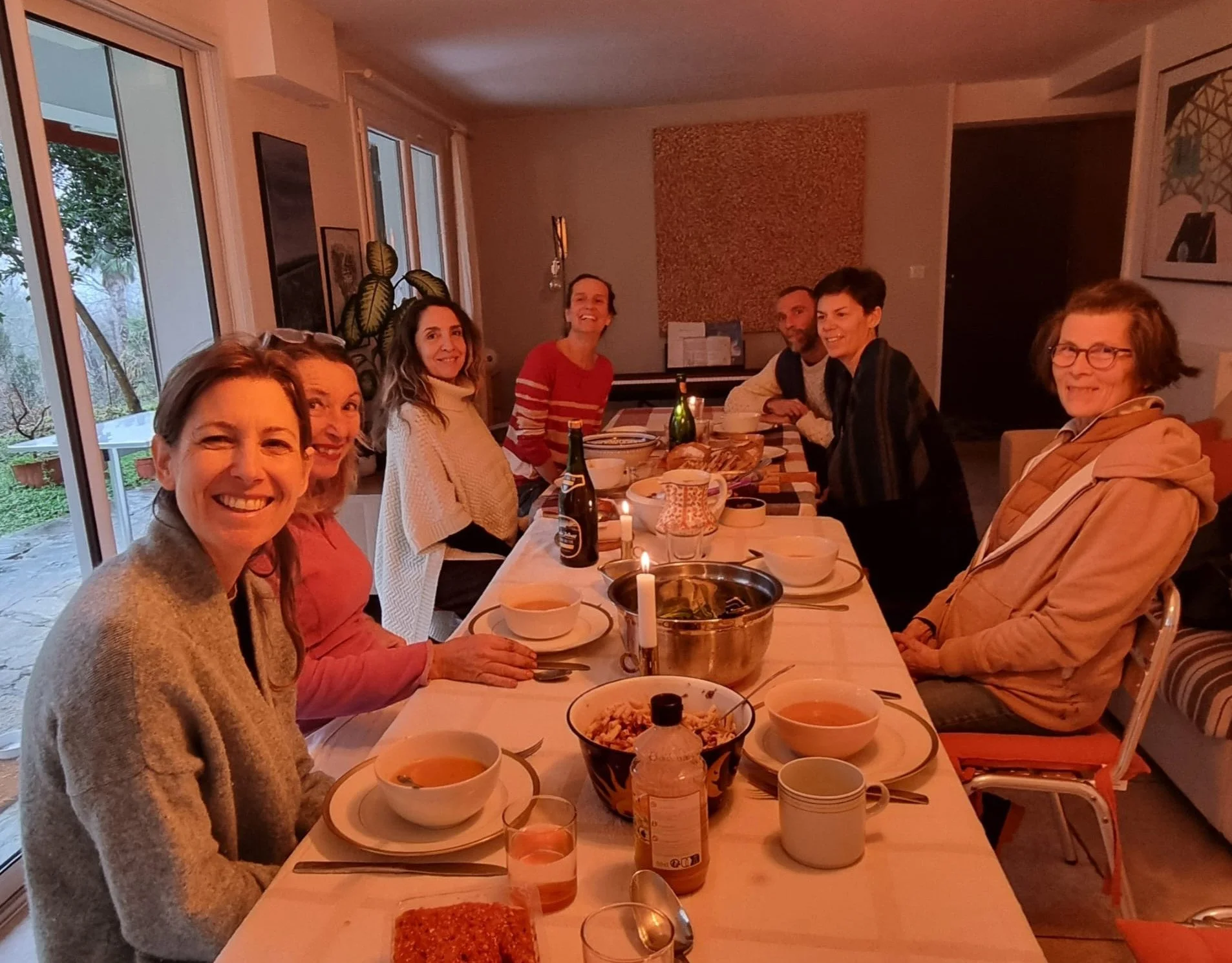 Groupe de huit personnes assises autour d'une grande table de dîner dans une maison, avec des assiettes, des bols, des boissons, et des bougies, à l'intérieur d'une pièce lumineuse avec des fenêtres et des décorations murales.
