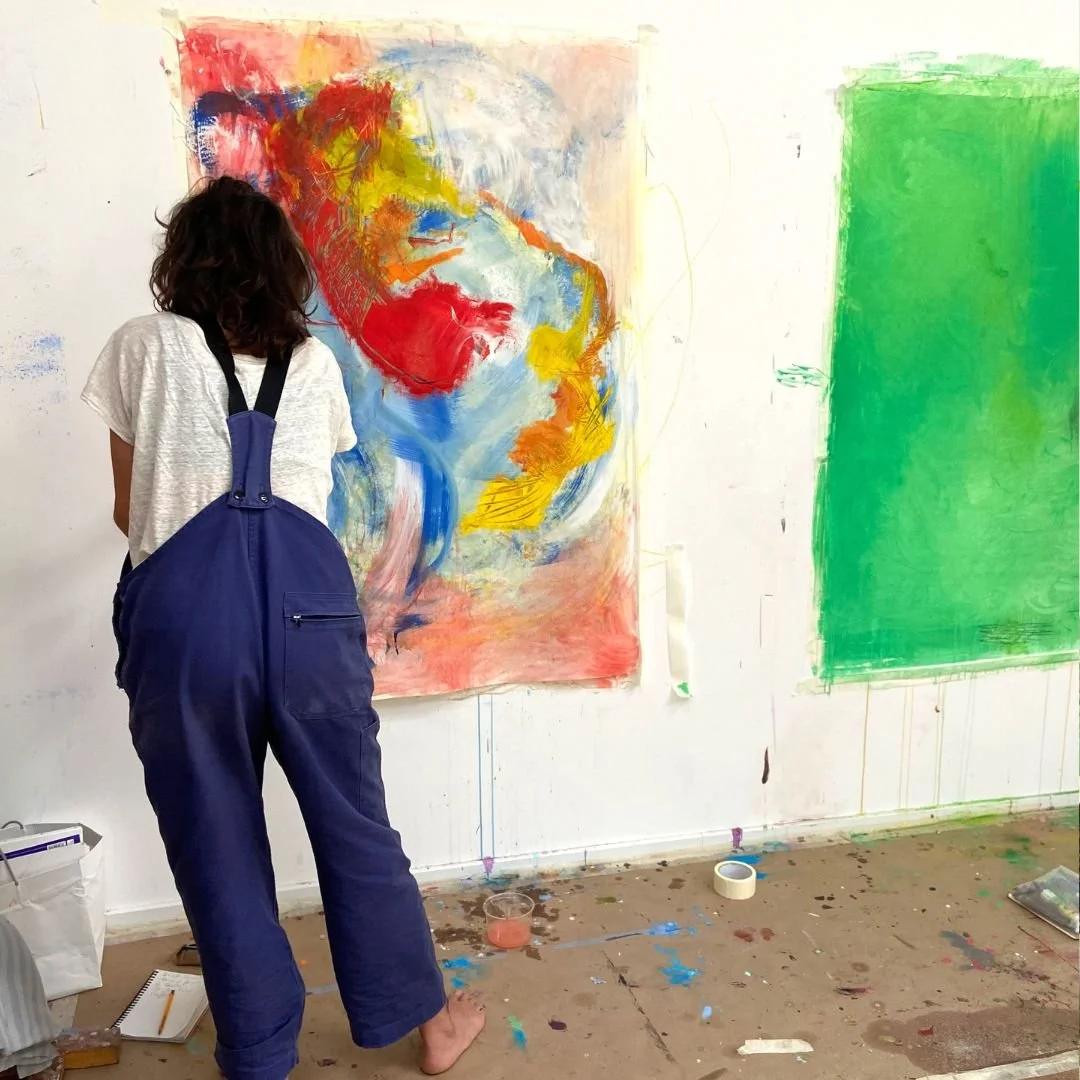 Une personne avec un overalls bleu, vue de dos, en train de peindre une toile abstraite colorée dans un studio d'art.