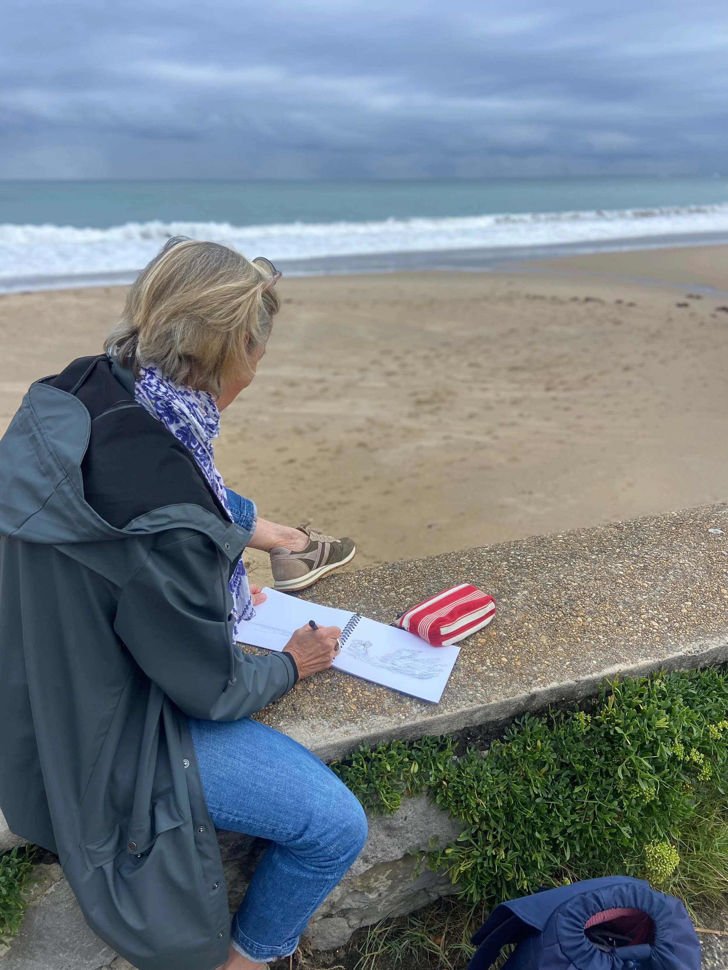 Une femme assise sur un banc au bord de la mer, dessinant dans un carnet. Elle porte une veste grise, des jeans bleus, et a des lunettes sur la tête. À côté d'elle, il y a un sac à dos rayé rouge et blanc posé sur le banc. Le ciel est nuageux, la plage est sablonneuse, et des vagues arrivent à la côte.