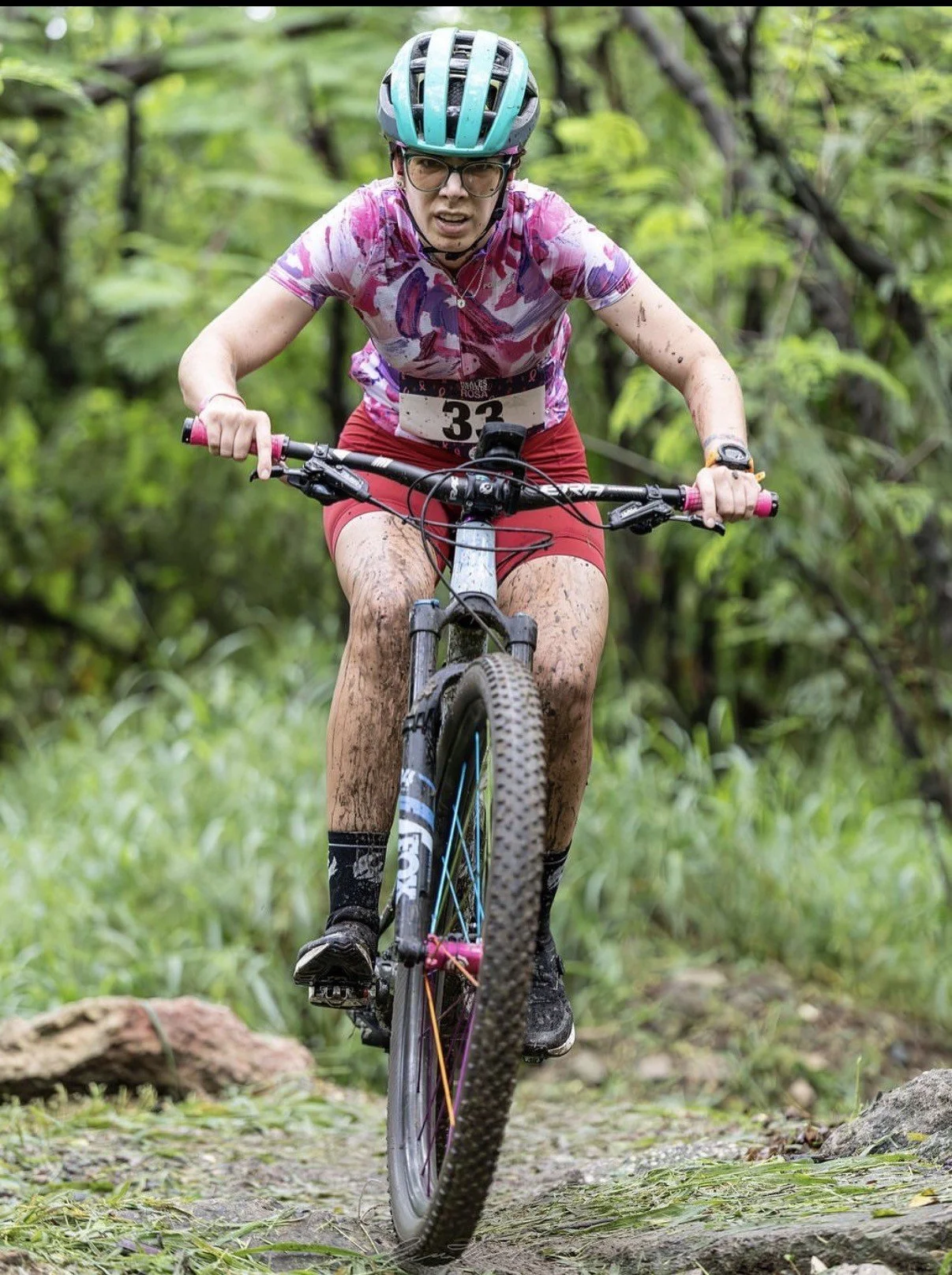 Una mujer andina en bicicleta de montaña en un sendero boscoso, vestida con casco, gafas, camiseta de color rosa con patrones y pantalones cortos. Está rodeada de vegetación.