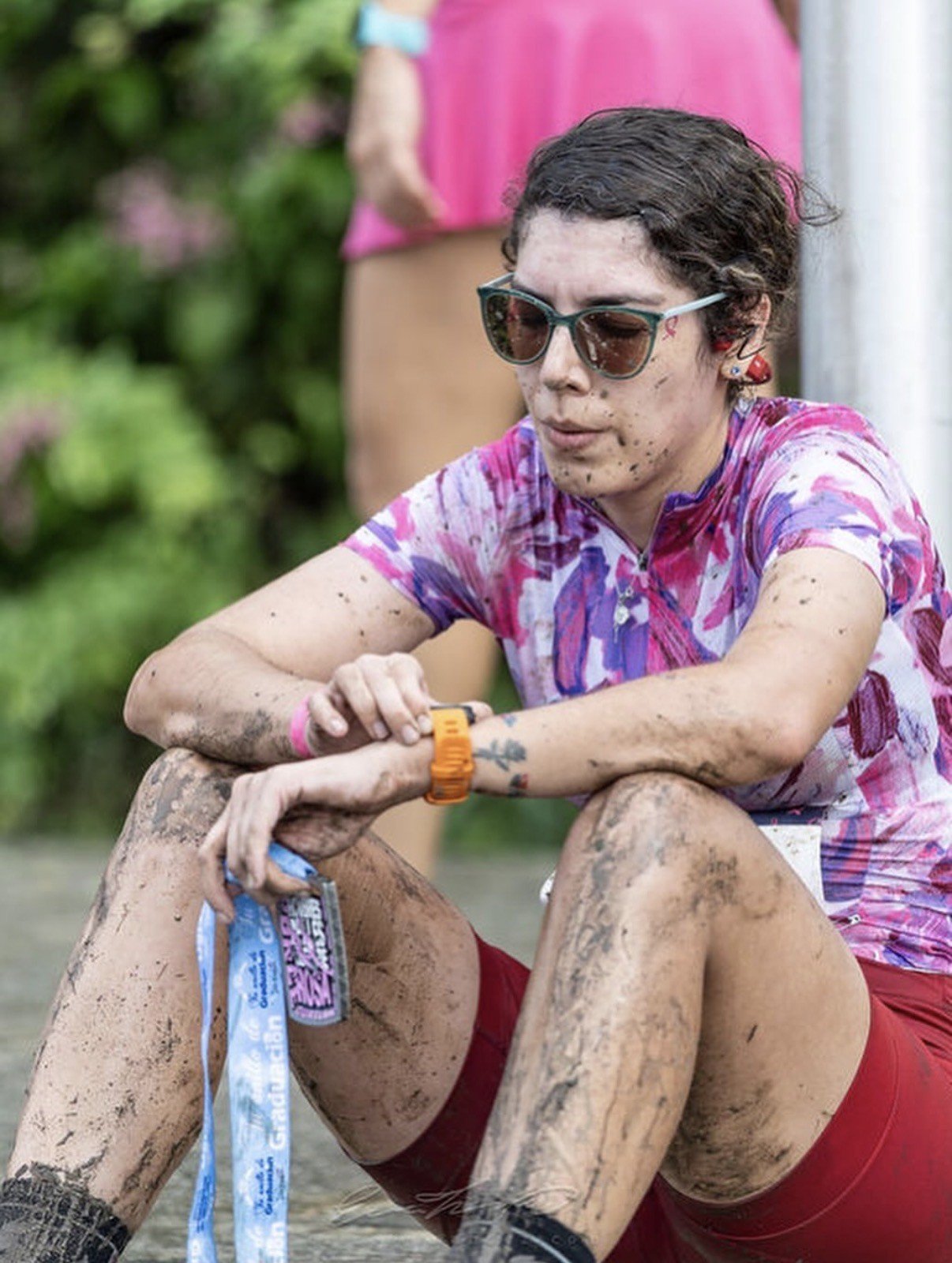 Personas en competencia de carrera atravesando una zona de barro, con una medalla en la mano y mostrando esfuerzo, vestidas con ropa colorida y accesorios como gafas de sol y reloj.
