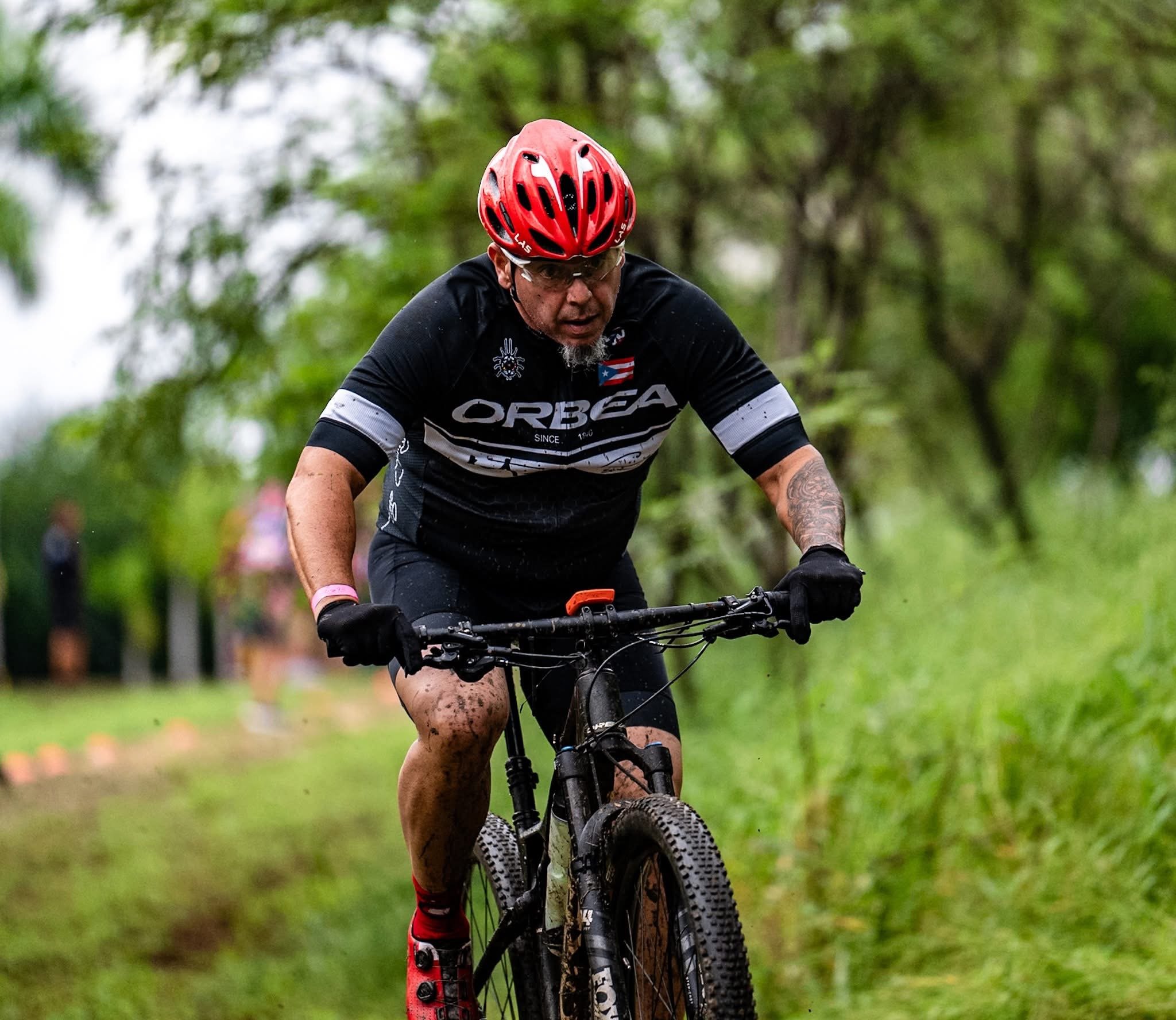 Un ciclista con casco rojo y camiseta negra, cubierto de barro, pedaleando en un sendero con fondo de árboles verdes.
