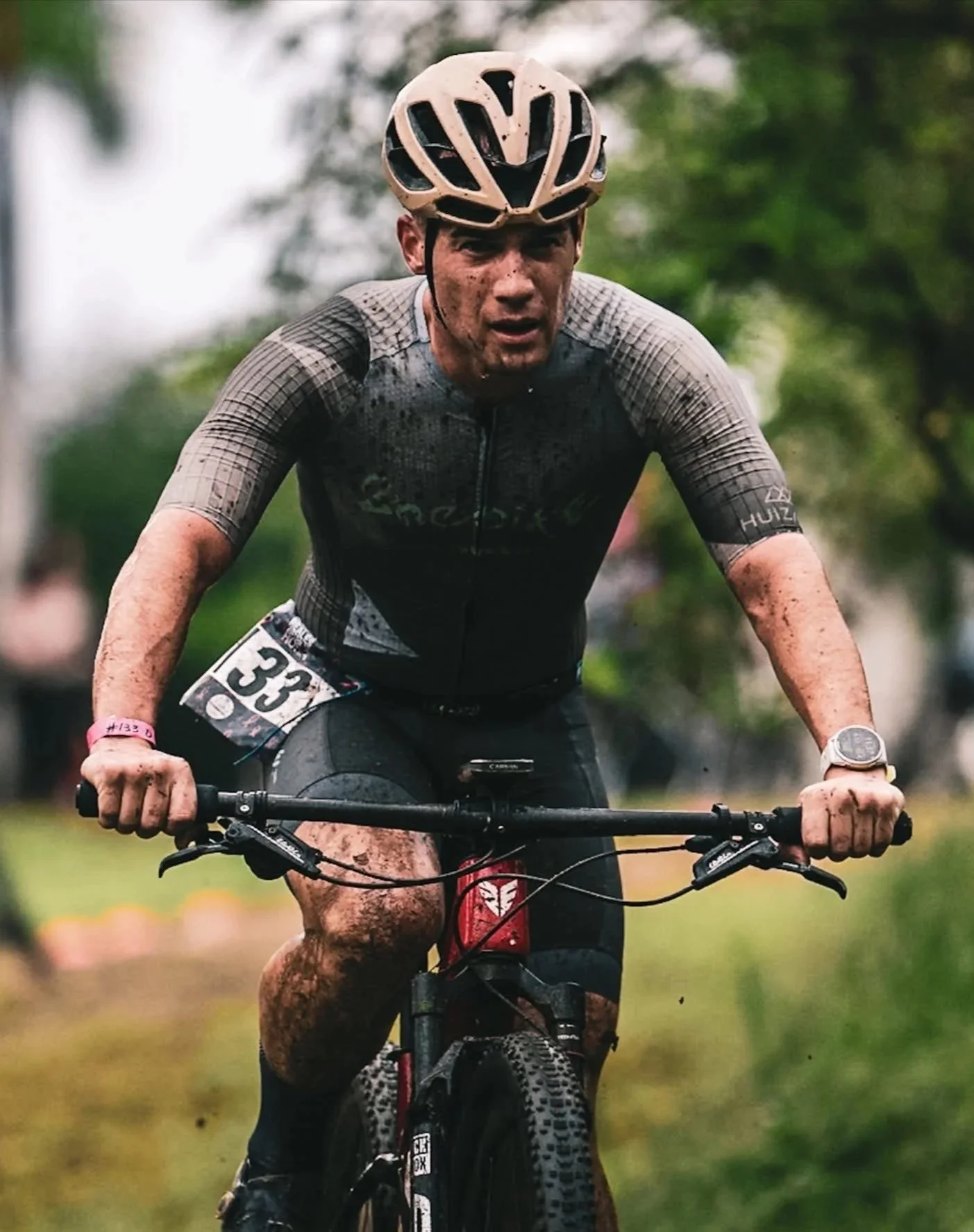Hombre en bicicleta de montaña con casco, ropa deportiva y número de competidor, pedalear en terreno embarrado en un evento de ciclismo de montaña.