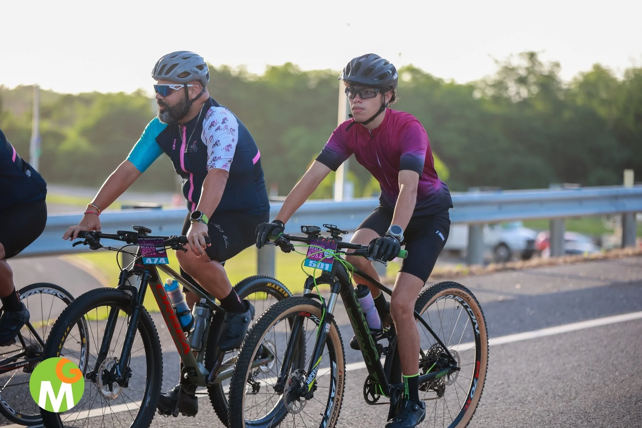 Dos ciclistas en bicicleta, uno con camiseta rosa y otro con camiseta de colores, con cascos y gafas, pedaleando en una vía al aire libre.