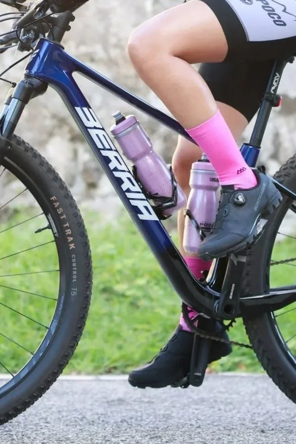 Primera mitad de una bicicleta de montaña azul marca Beruja, con un marco robusto, y dos botellas de agua rosadas en los portabotellas. Un ciclista, con una pierna desarrollada y vestimenta en negro, lleva calcetas rosadas, un zapato negro y una parte de su ropa deportiva. La bicicleta está en una superficie pavimentada con césped verde y fondo difuso de rocas o pared de piedra.