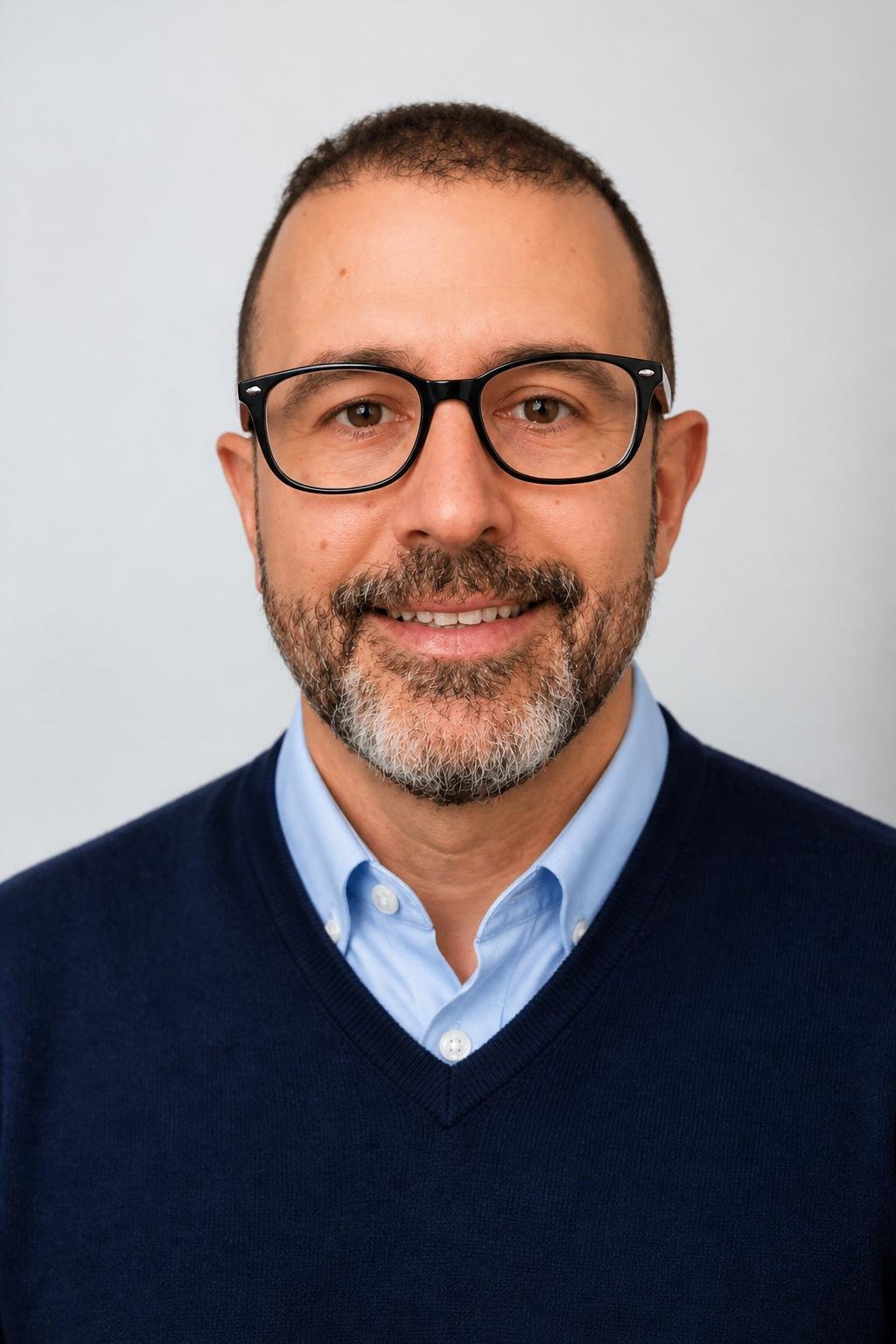 Close-up of a middle-aged man with glasses, a beard, wearing a light blue collared shirt and a dark blue sweater, smiling against a plain background.