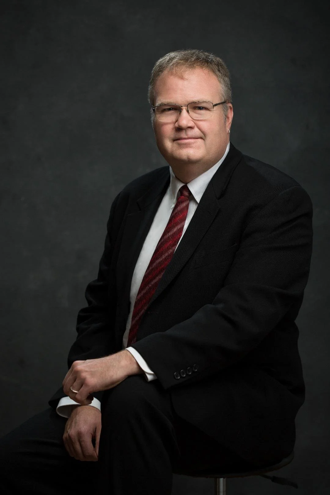 Portrait of a man wearing glasses, a black suit, white shirt, and red tie, seated against a dark background.