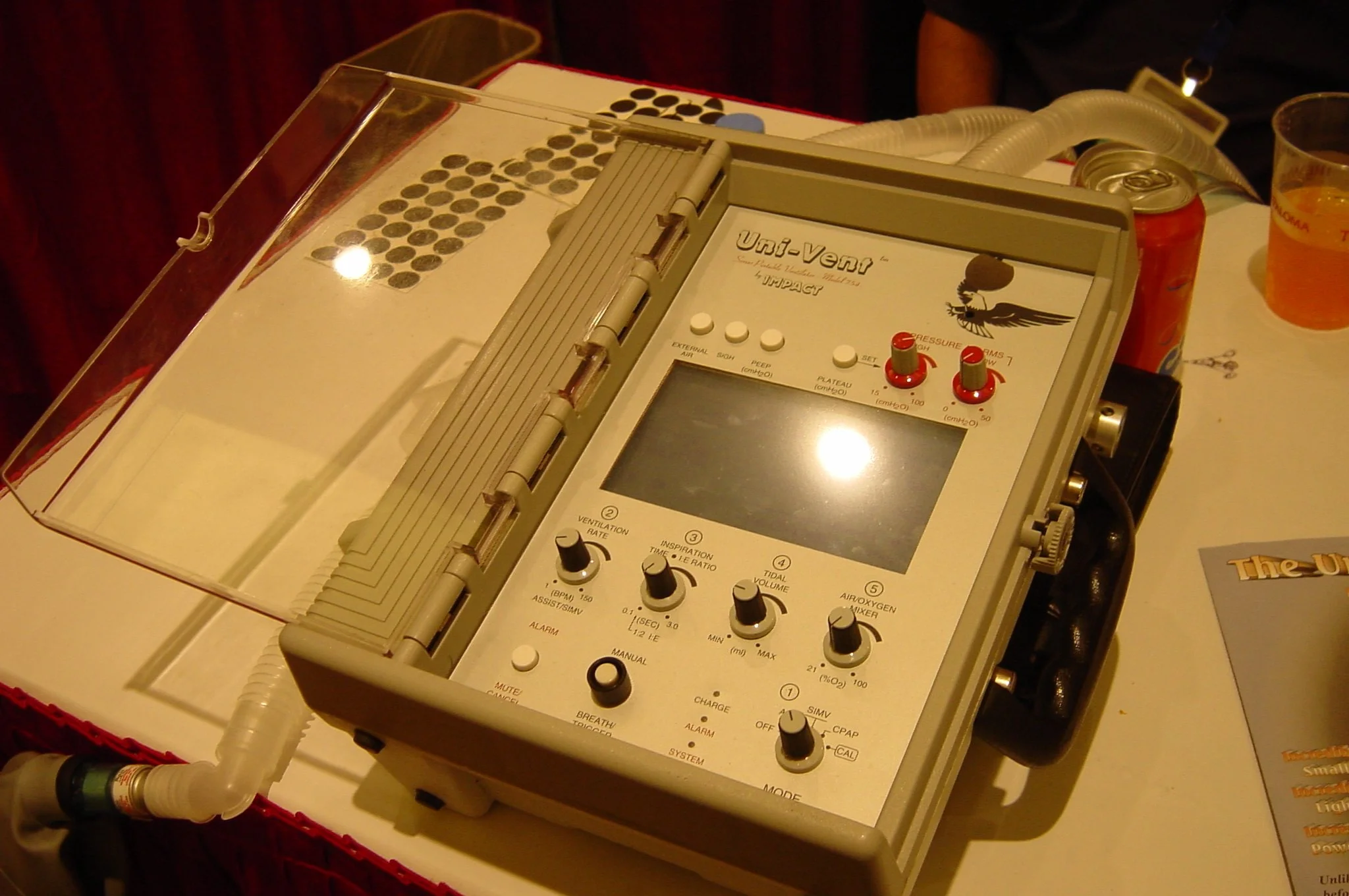 Medical ventilator with control knobs and tubing on a table, next to drinks and a brochure.