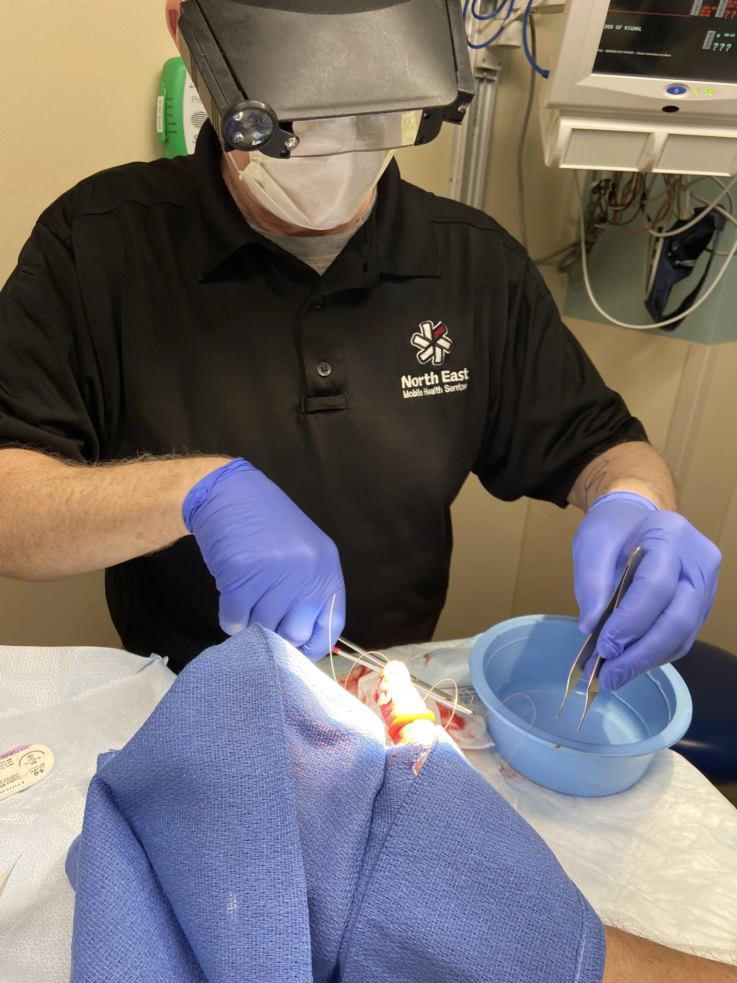 A medical professional wearing protective gear and gloves performing a surgical procedure on a patient. The healthcare worker is looking down at the patient, with medical equipment visible in the background.