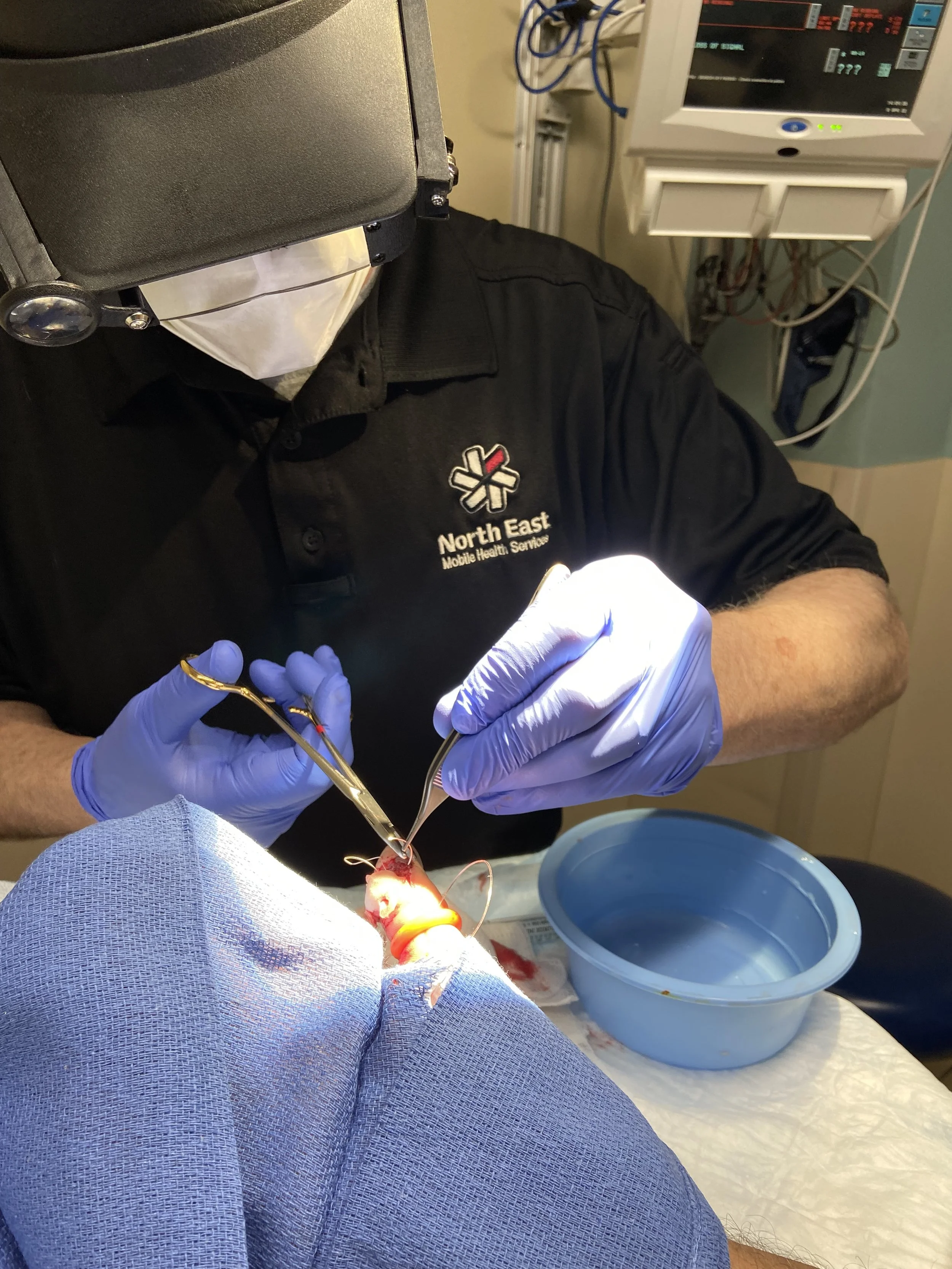 A medical professional wearing blue gloves and a black shirt with 'North East Mobile Health Services' is performing a surgical procedure on a patient, who is covered with a blue cloth. Medical equipment, monitors, and wires are visible in the background.