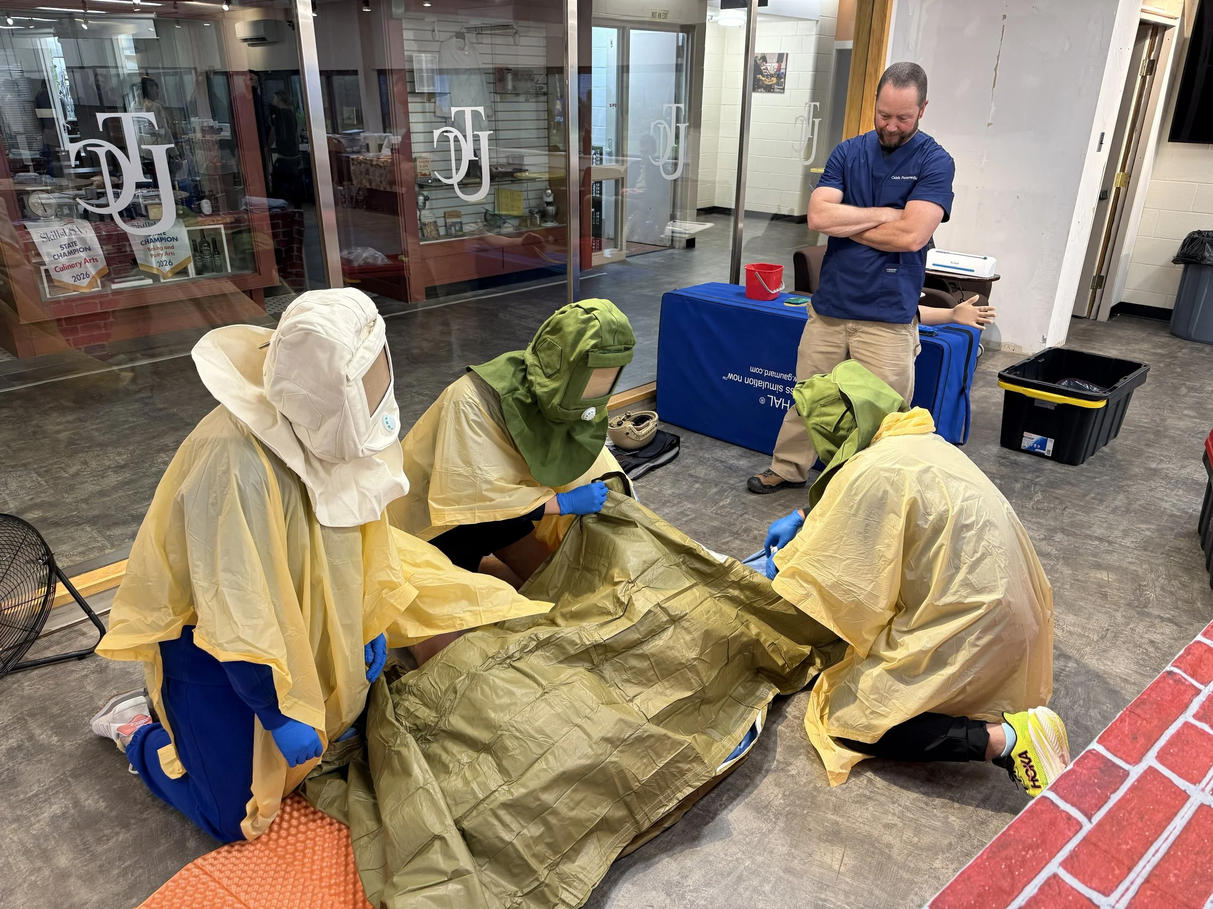 A group of people dressed in protective gear, including yellow coveralls, blue gloves, and masks, are gathered on the floor of an indoor space. They appear to be participating in a training or medical emergency drill, with a man in a blue shirt supervising or instructing them. The room has a glass wall with signs and awards displayed on the other side.