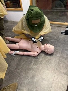Person wearing a green cap and yellow apron practicing CPR on a mannequin in a classroom or training environment.