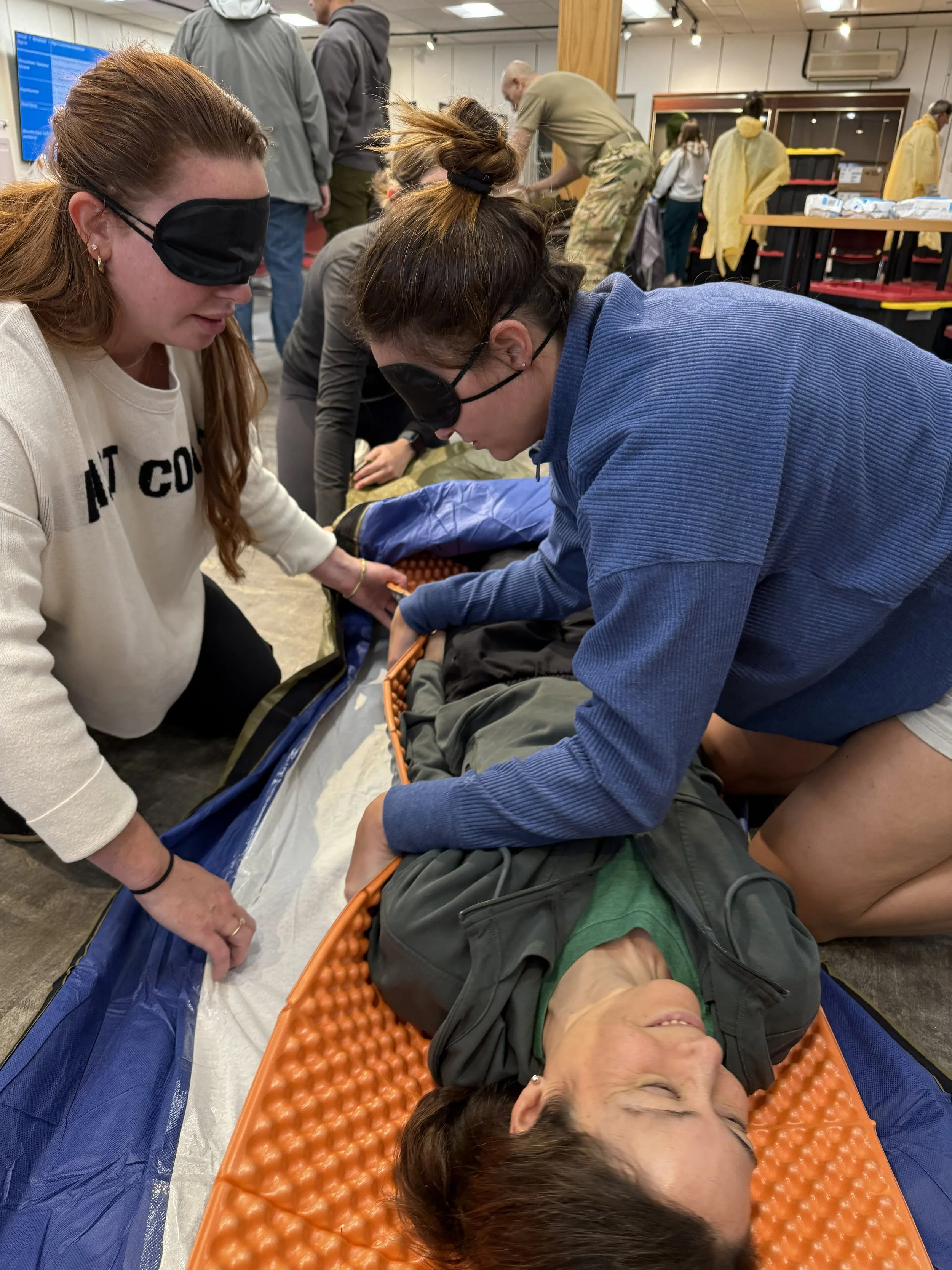 People participating in a CPR training session, practicing on a person lying on a padded mat, with others in the background observing.