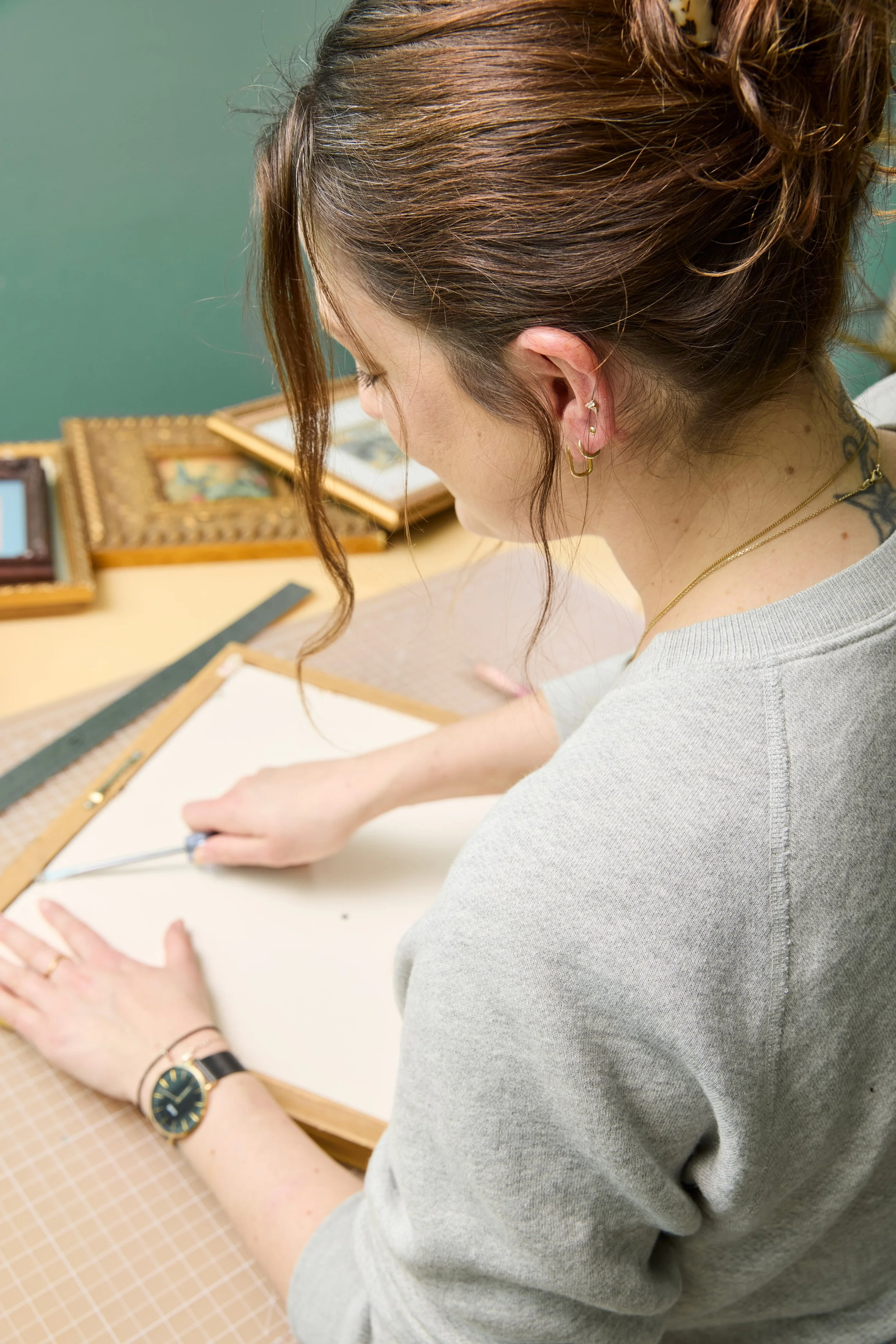 A woman with brown hair styled in a loose updo is working on a craft project at a table, with several framed pictures in the background.
