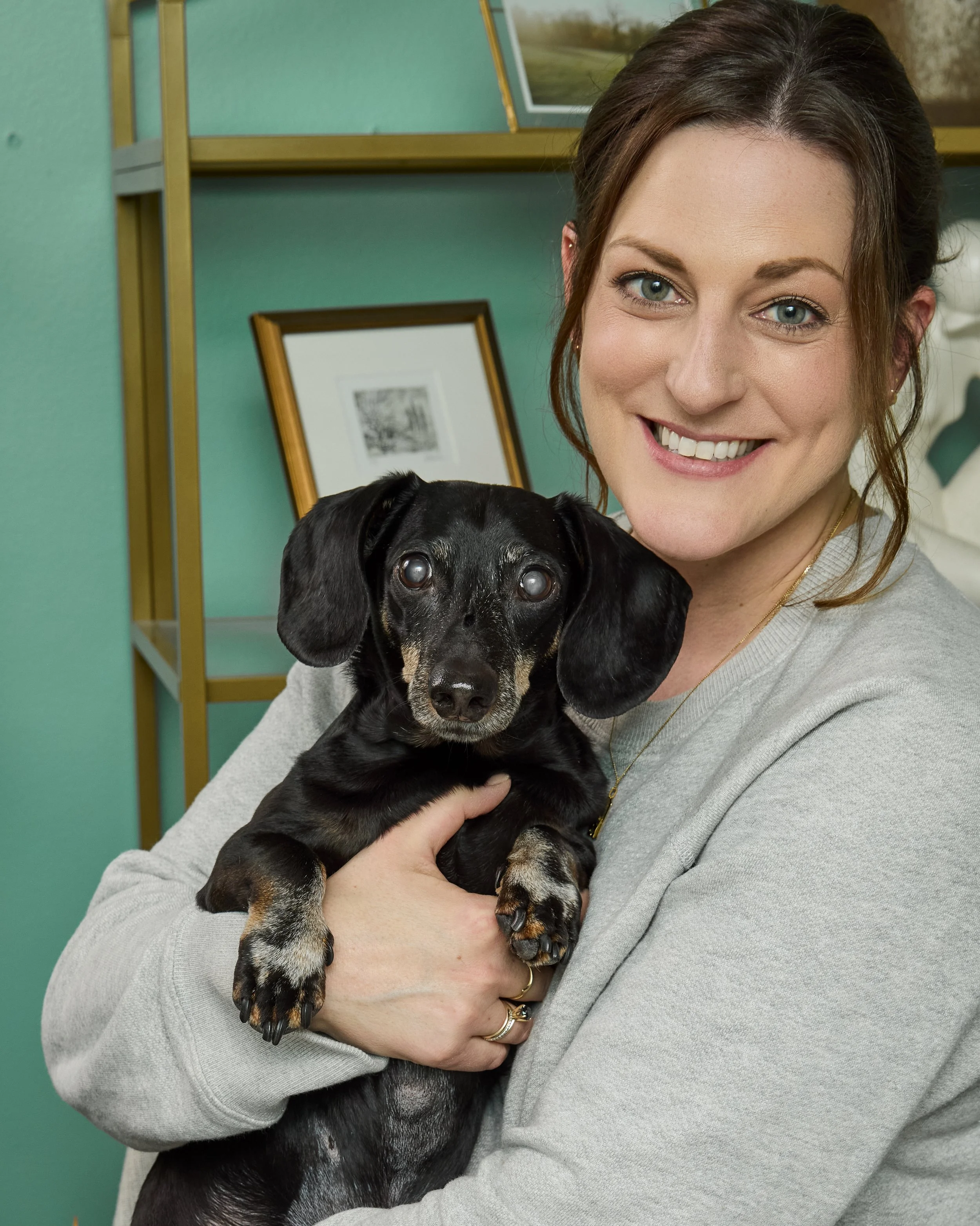 A woman with brown hair and green eyes smiling and holding a black and tan puppy, in an indoor setting with teal walls and framed pictures.