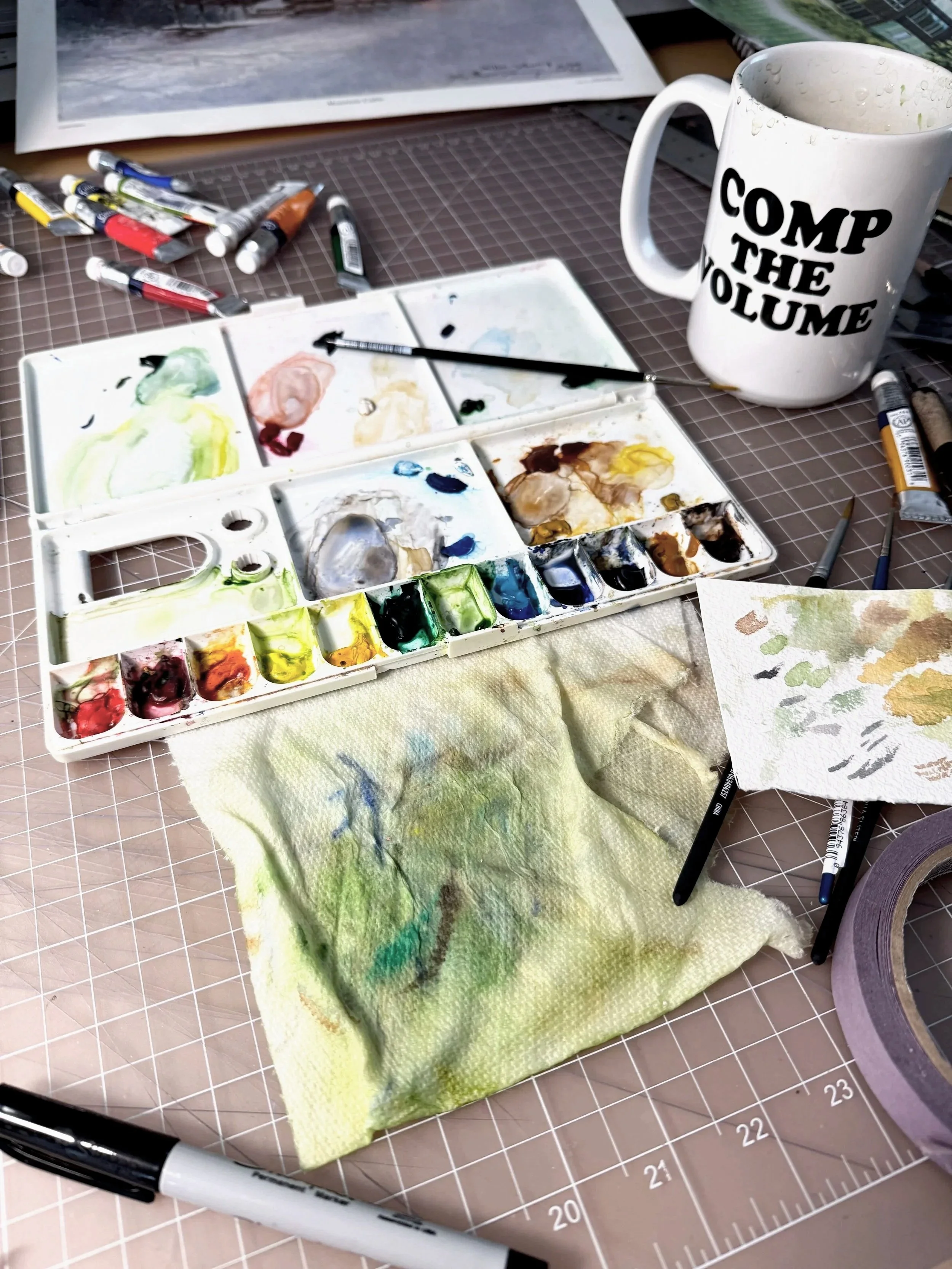Watercolor paint palette with mixed paint colors, paintbrush, piece of watercolor paper with color swatches, a white mug with black text, pens, and a paper towel with watercolor stains on a cutting mat.