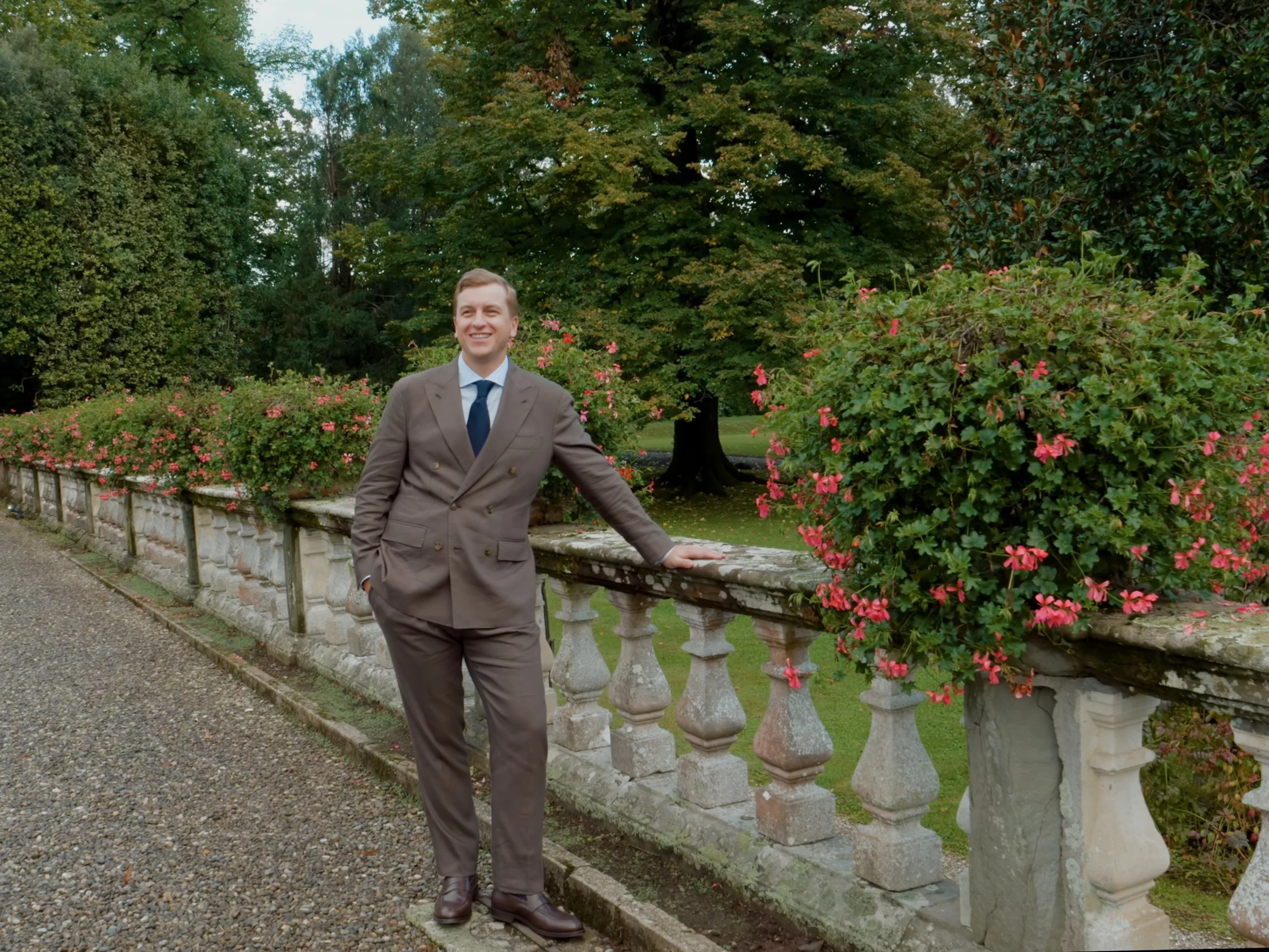 A man in a brown suit, white shirt, and tie standing outdoors near a stone and concrete balustrade with pink flowers and green trees in the background.